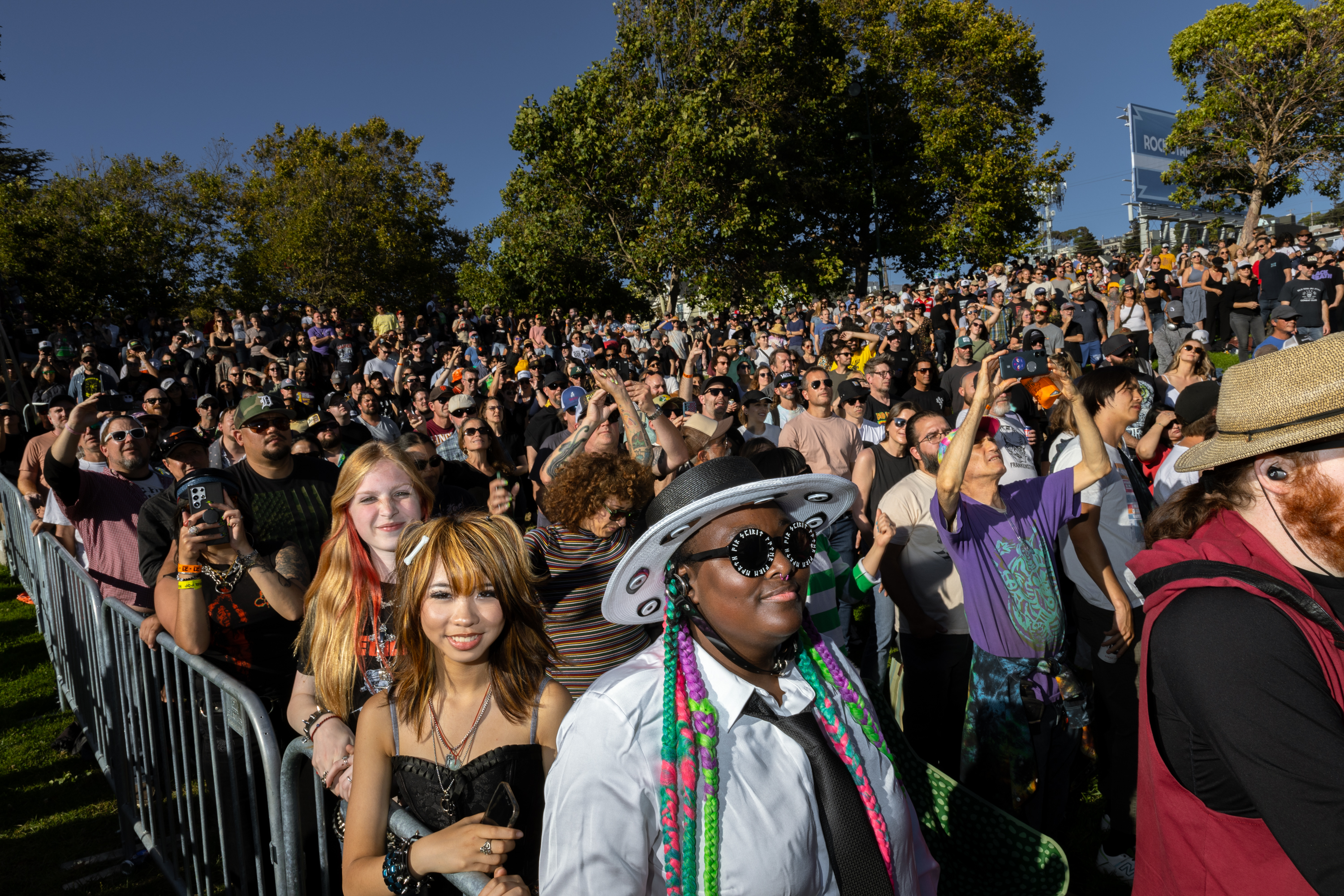 A diverse crowd of excited people enjoy an outdoor event, many smiling and taking photos, with trees and a clear sky in the background.