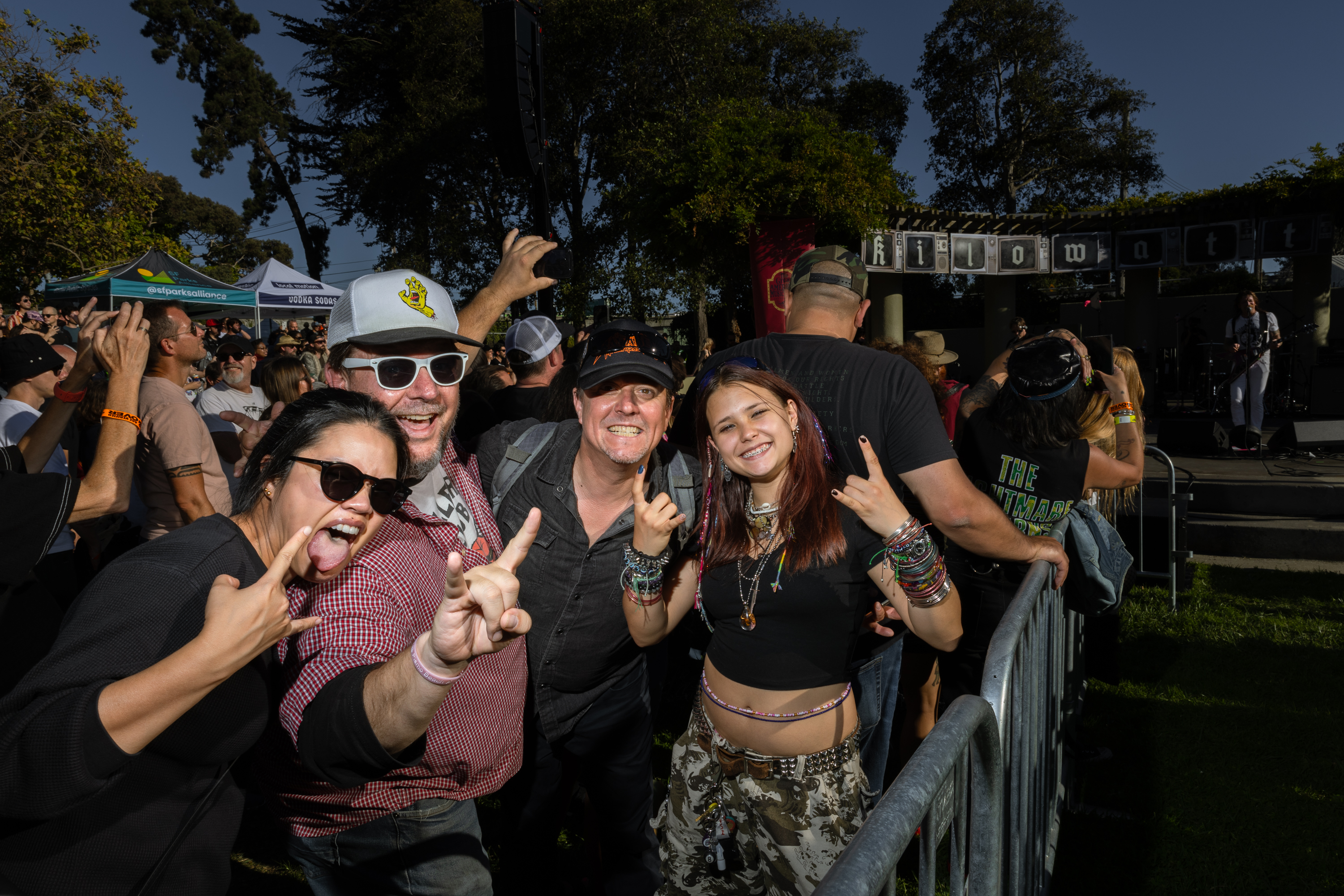 Four people at an outdoor concert smile, make hand gestures, and seem excited. They are surrounded by a crowd near a stage with a performing band.