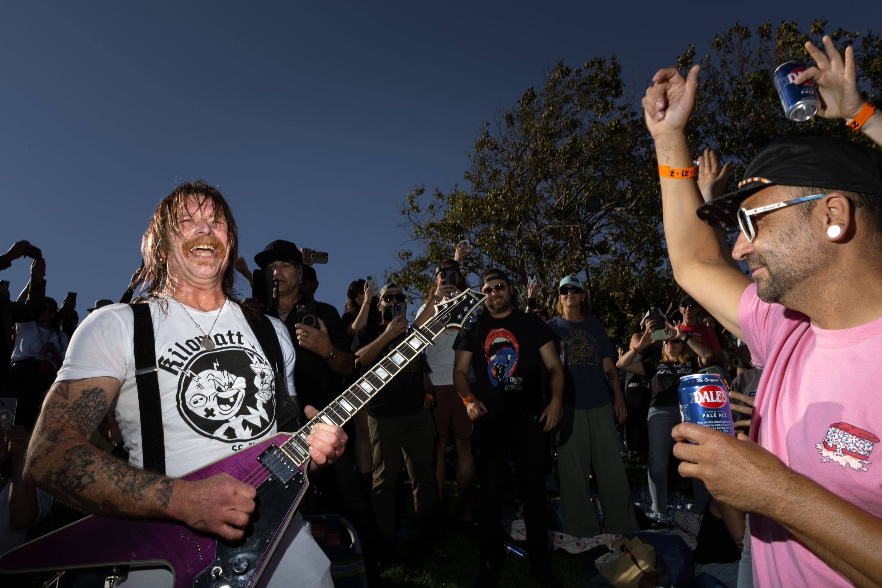 A man with long hair and tattoos plays a purple electric guitar joyfully amid a lively, cheering outdoor crowd; one person is raising his arm with a beer can.