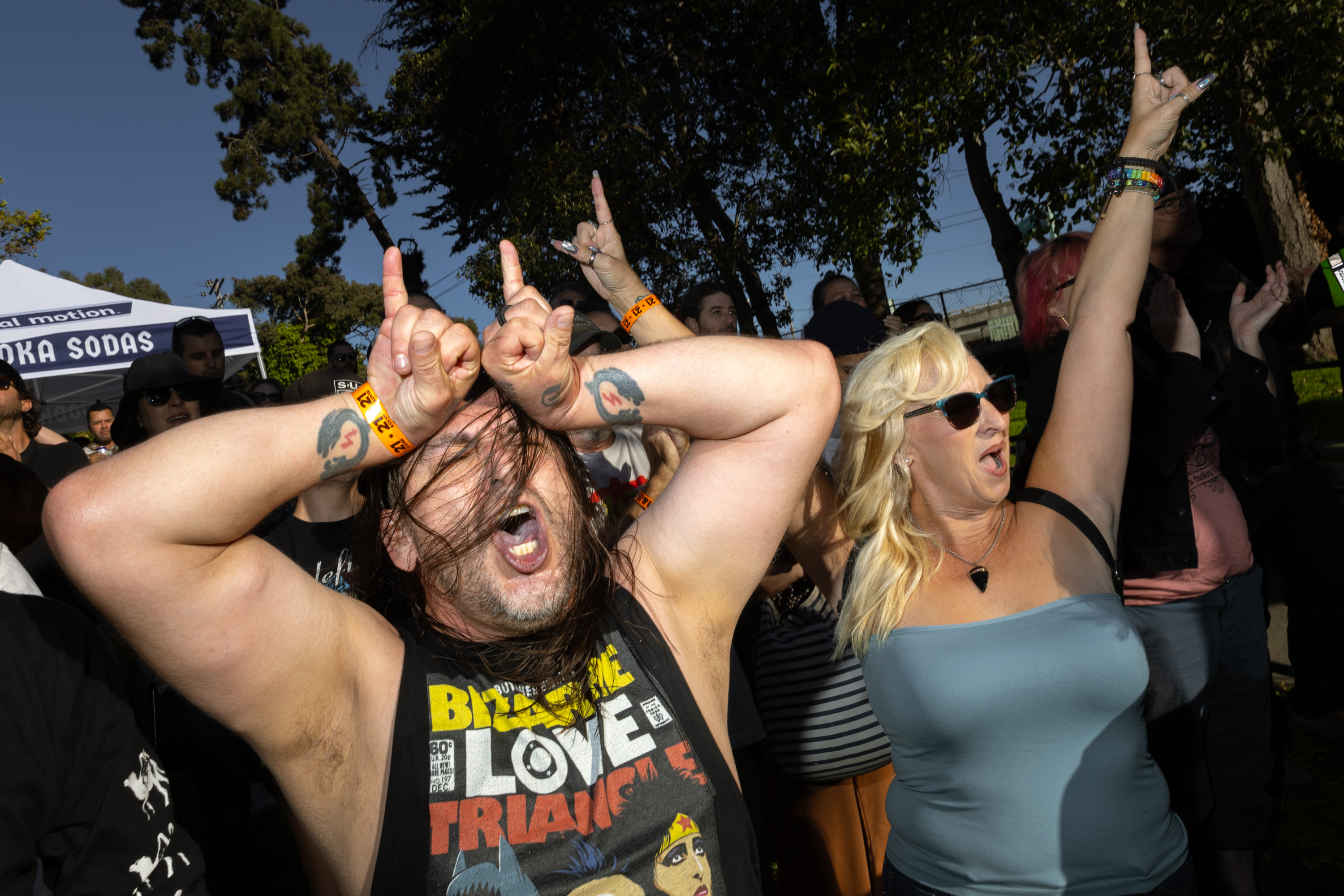 Two people at an outdoor event raise their hands making rock and roll horns gestures. The man has tattoos, a graphic tank top, and the woman wears sunglasses. They look excited.