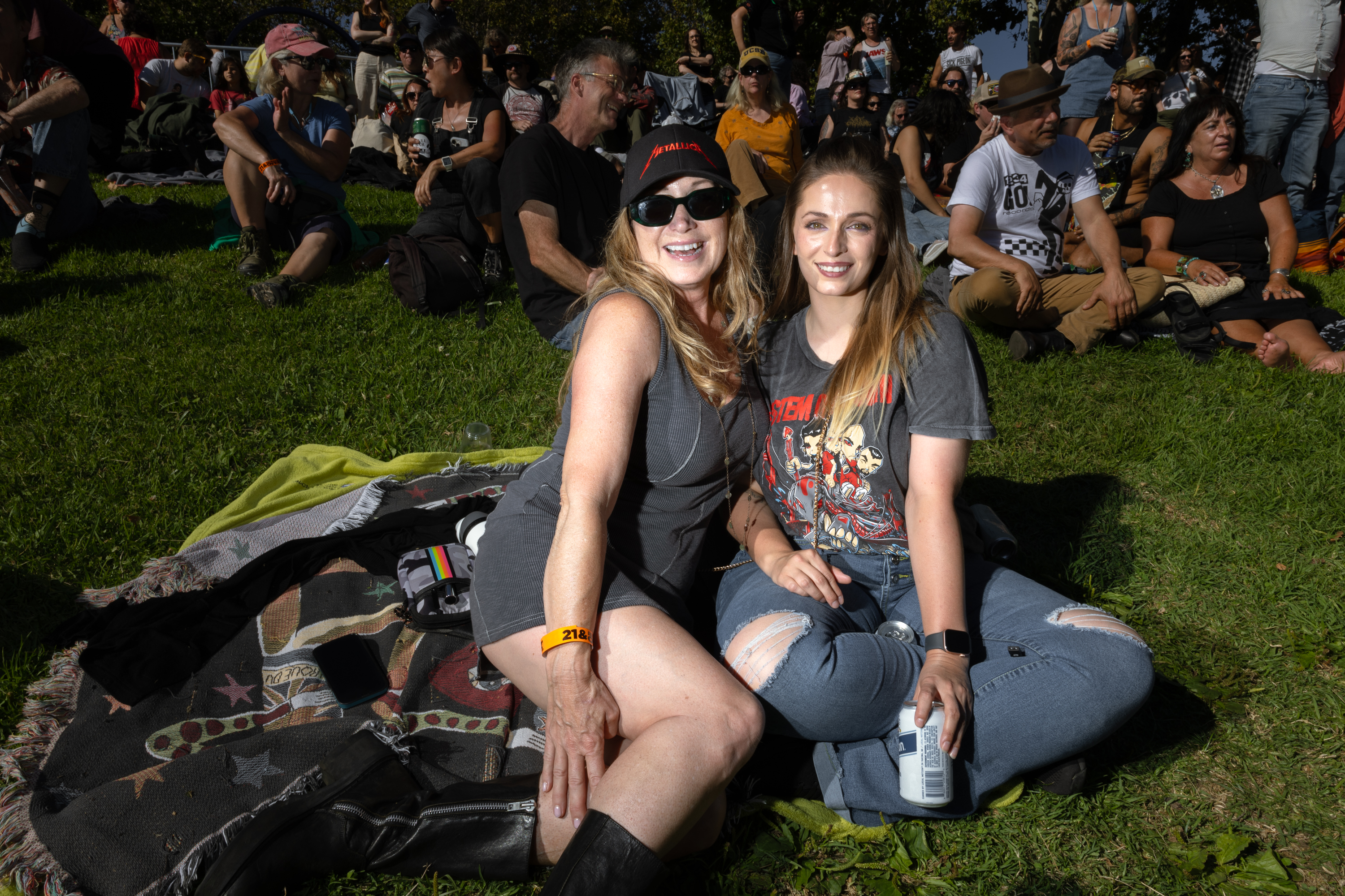 Two women are sitting on a blanket in a park, smiling at the camera. They are surrounded by other people sitting and talking on the grass, enjoying a sunny day.