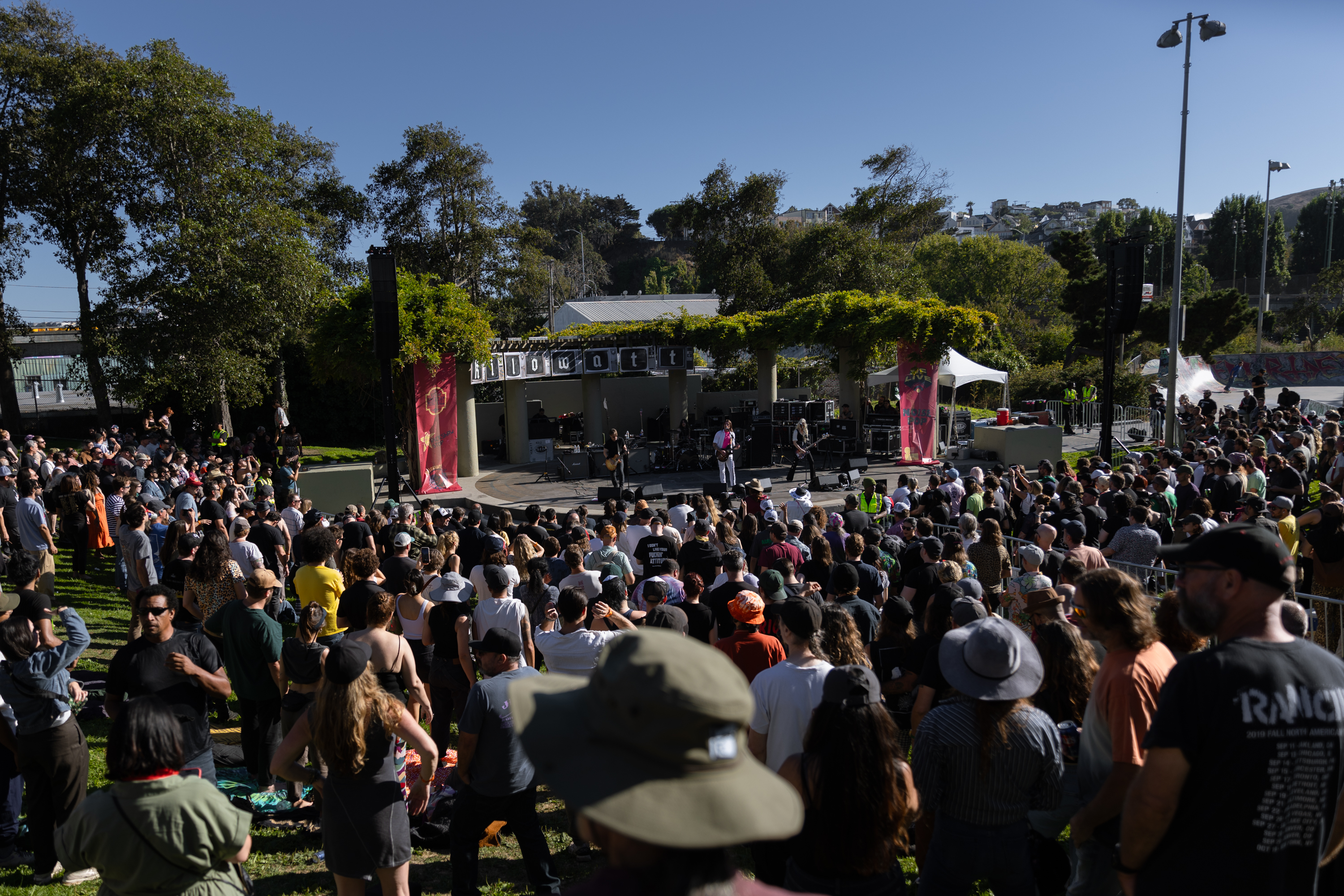 A large crowd gathers in a park on a sunny day, watching a band perform on an outdoor stage decorated with banners. There are trees and hills in the background.