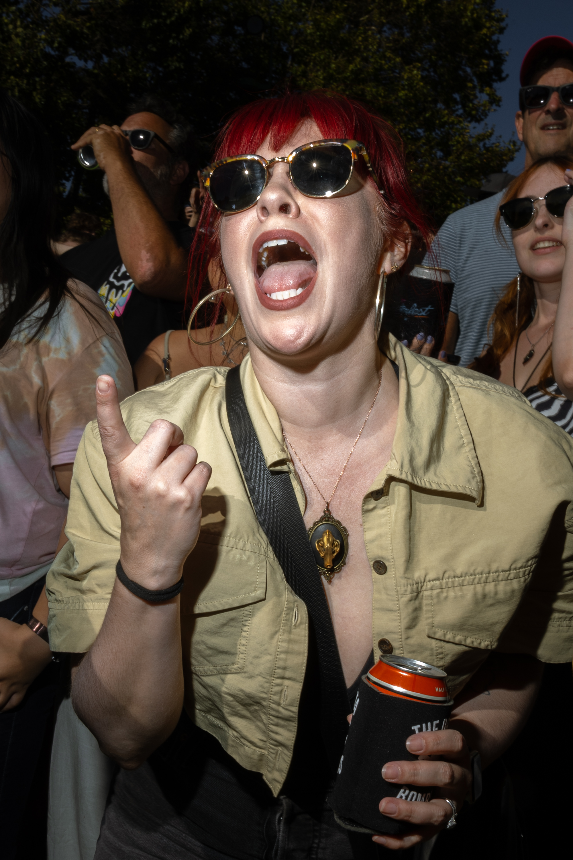 A woman with red hair and sunglasses enthusiastically shouts while holding a drink can. She wears a beige shirt, large hoop earrings, and a pendant necklace. People are around her.