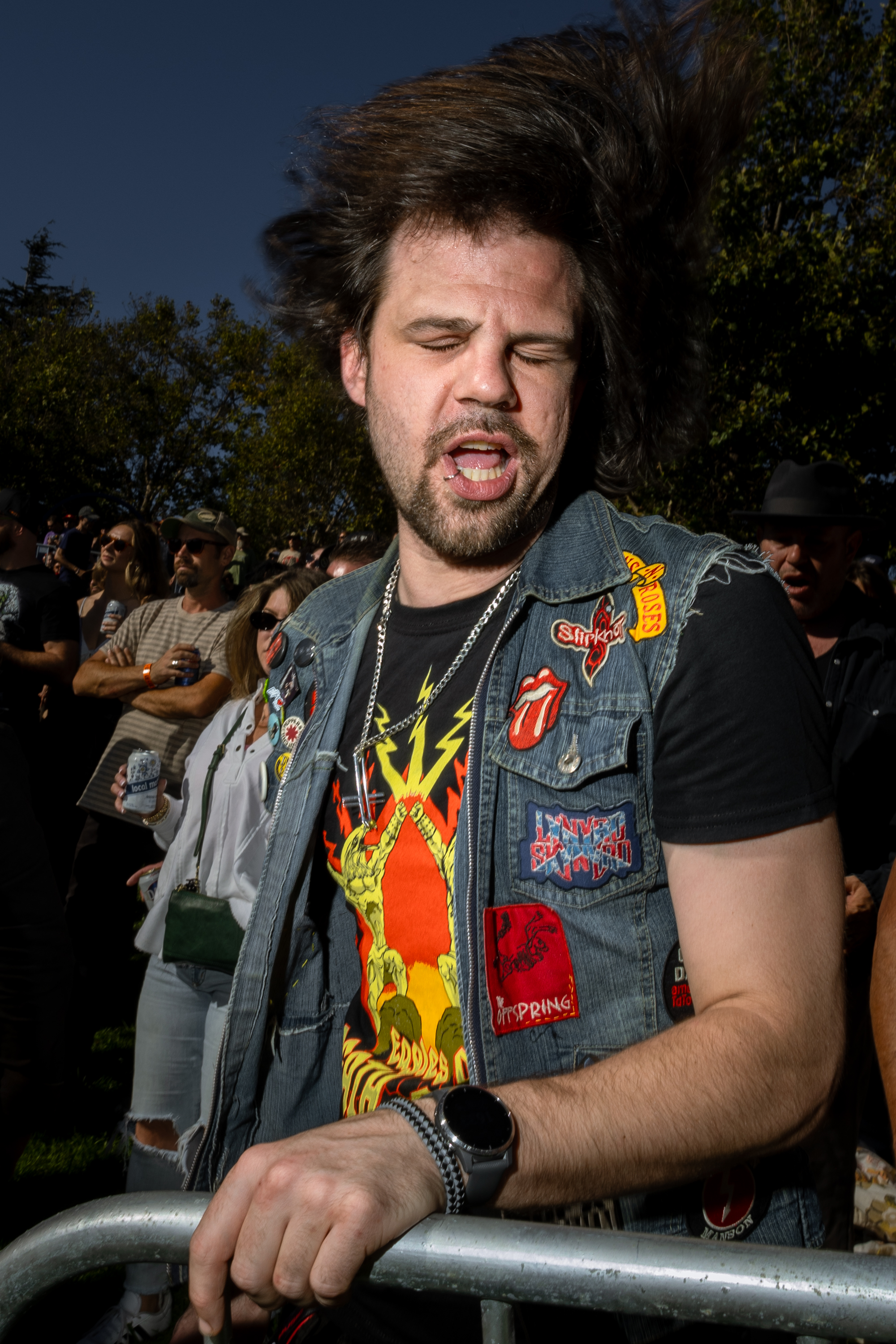 A man with wavy hair flips his head back, eyes closed, wearing a denim vest with music patches, and a graphic t-shirt, standing at an outdoor event with a crowd behind him.