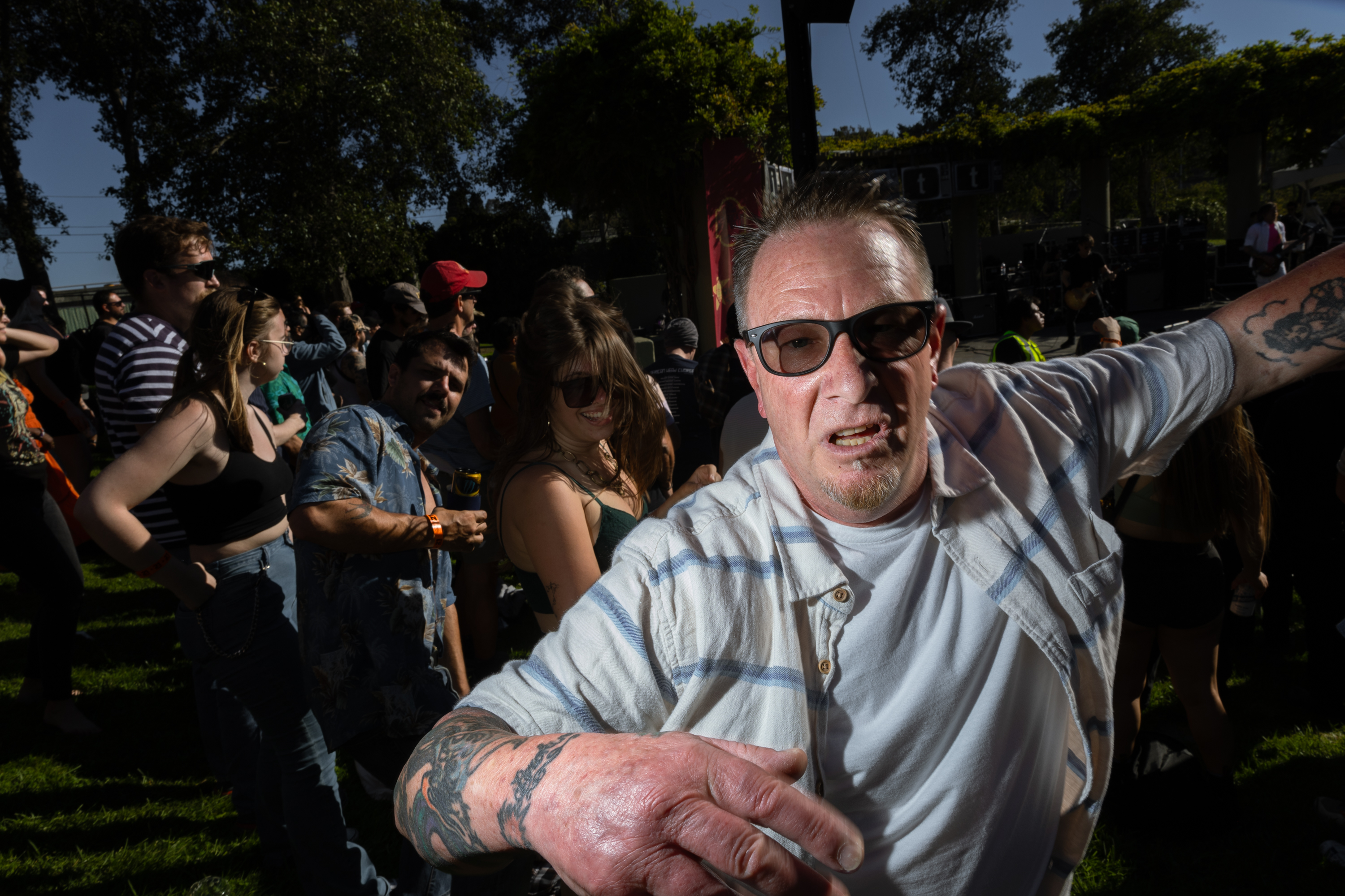 A visually striking man with glasses and tattoos leans into the camera, while a crowd of people, including some dancing and others watching a stage, surrounds him in an outdoor setting.