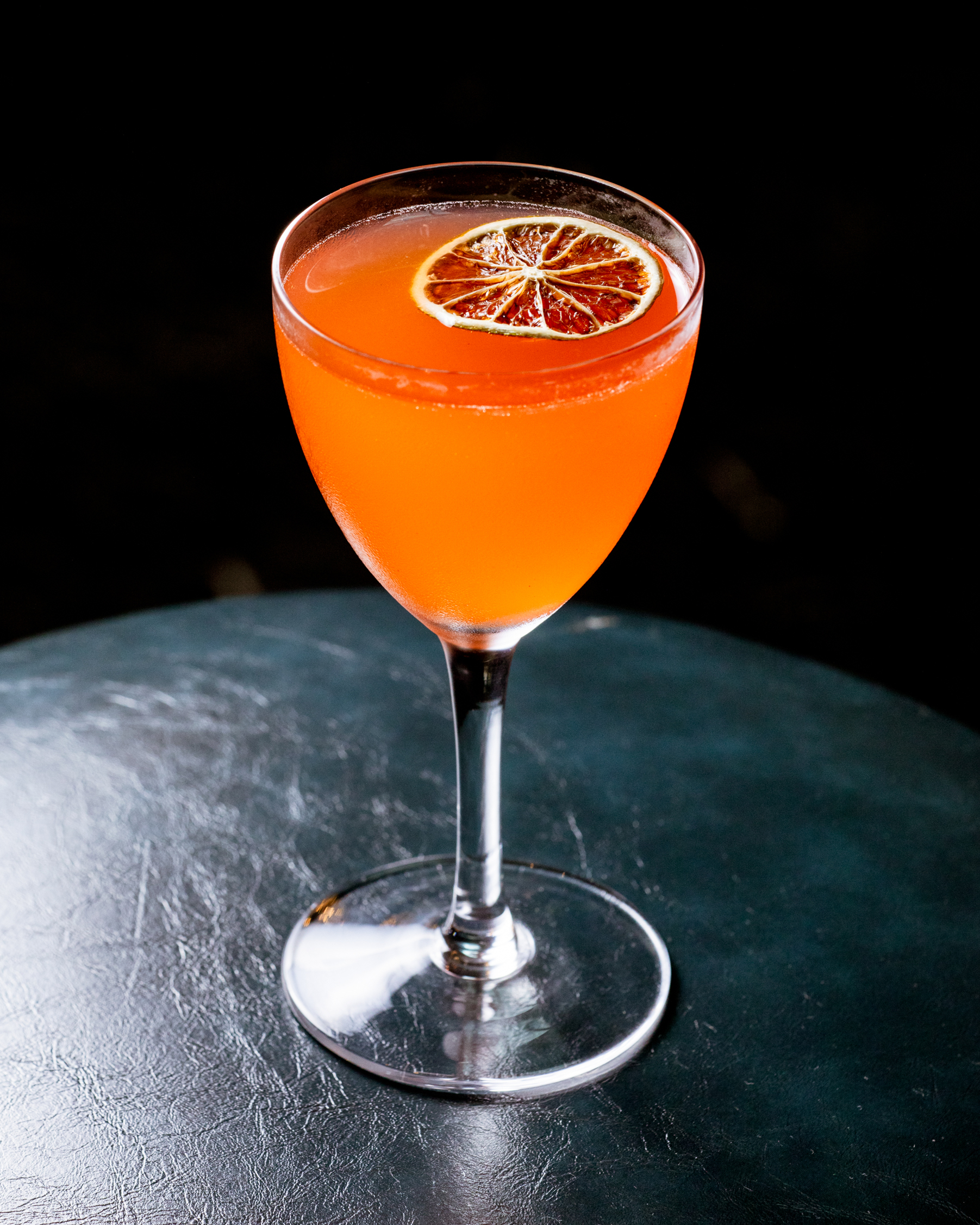 An orange cocktail in a stemmed glass garnished with a dried citrus slice sits on a dark, textured surface against a black background.