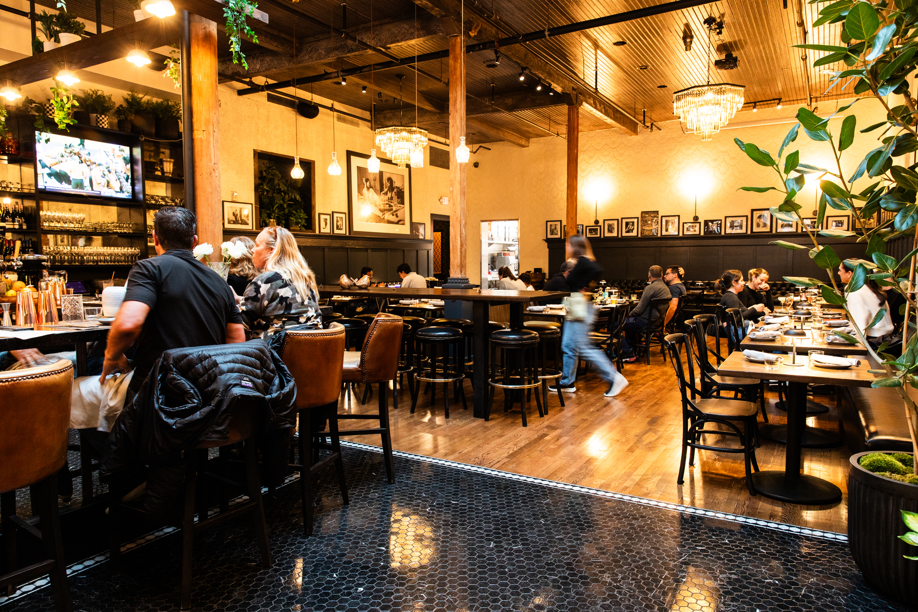 The image shows a busy restaurant with people dining and a bar area with patrons seated on stools. The decor is warm with wooden beams, hanging lights, and framed photos.