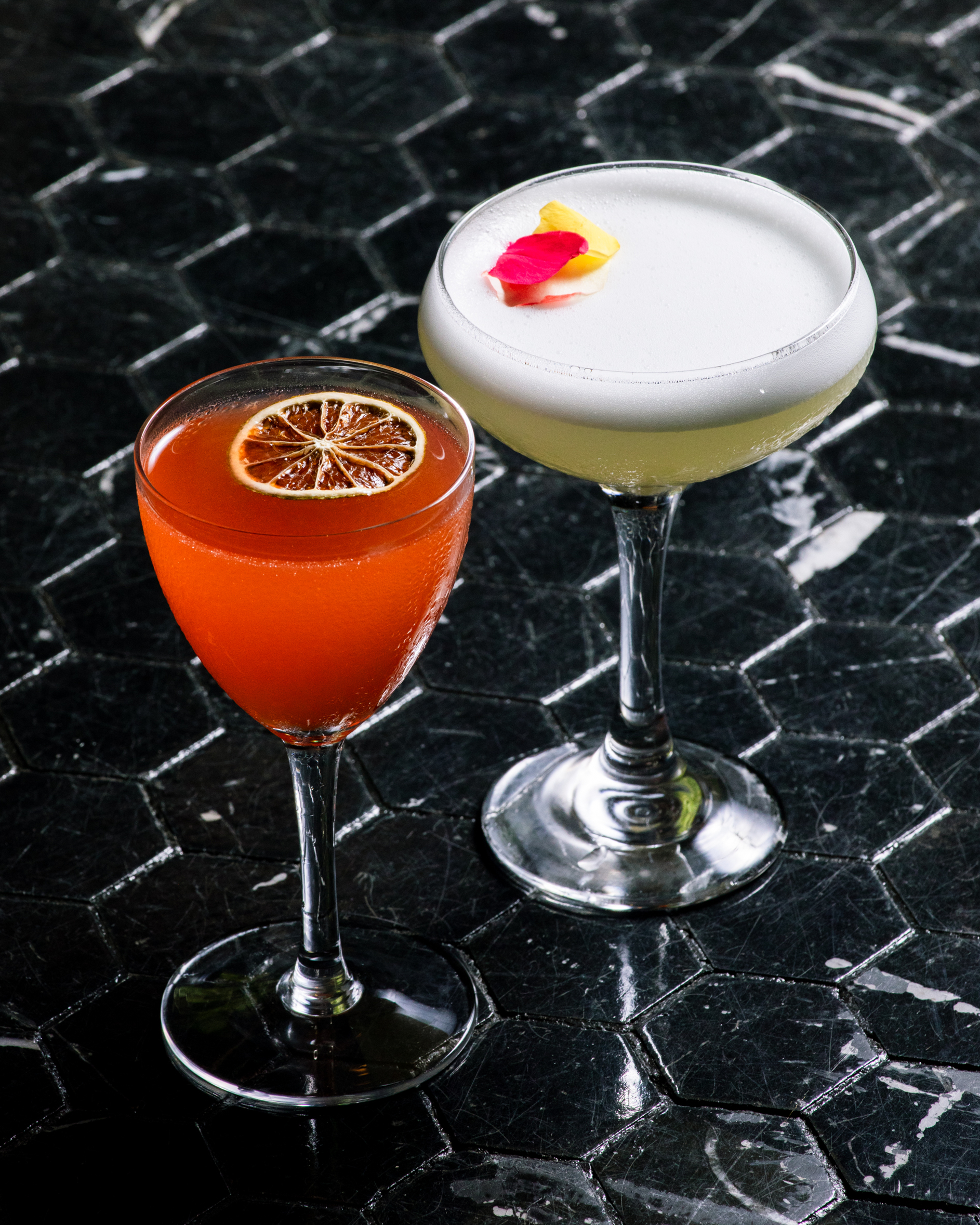 Two cocktails on a dark, hexagonal-tiled surface: one orange with a dried lime slice, the other pale yellow with a frothy top and flower petals.