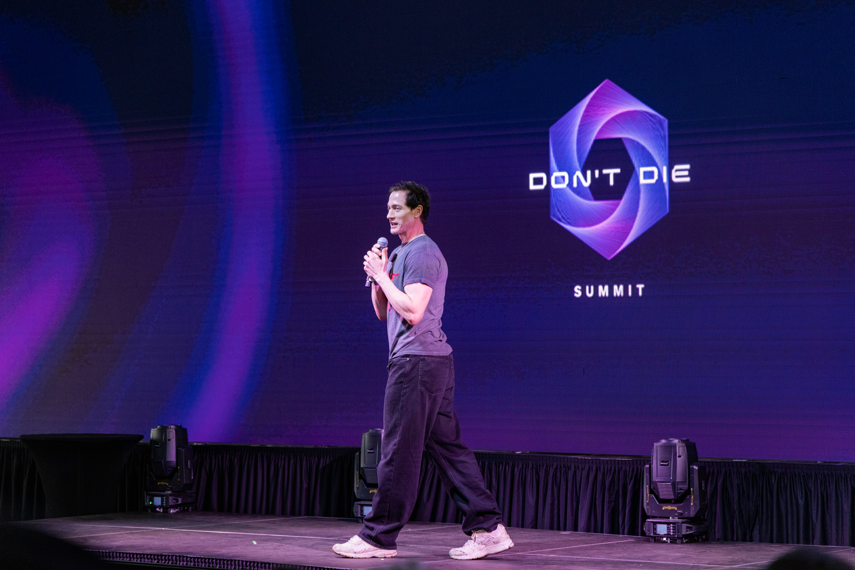 A man holds a microphone while standing on stage, speaking at the "Don't Die Summit," with a colorful geometric logo displayed on a large screen behind him.