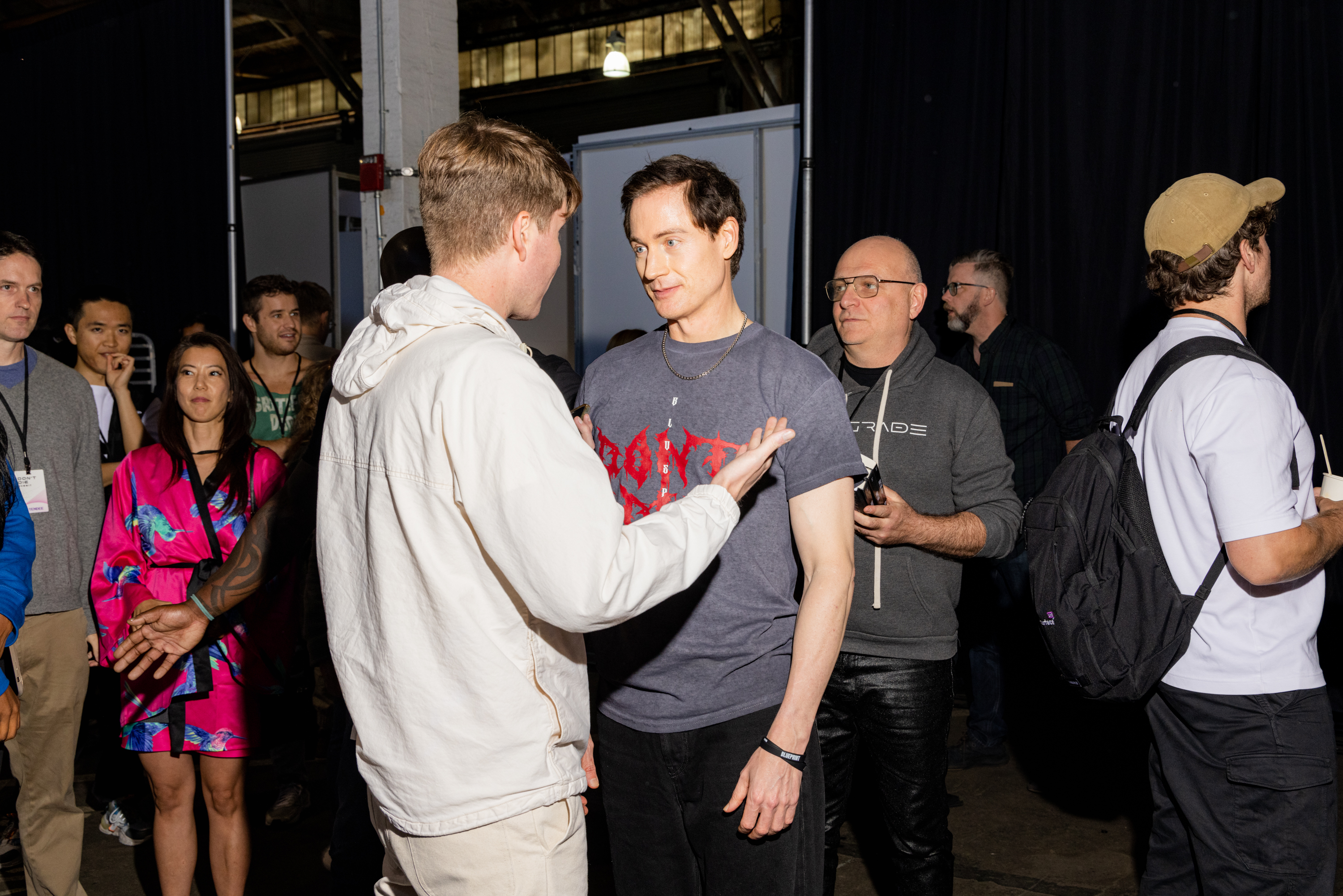 Several people are gathered indoors; two men in the foreground are engaged in conversation, while others in the background are chatting or observing.