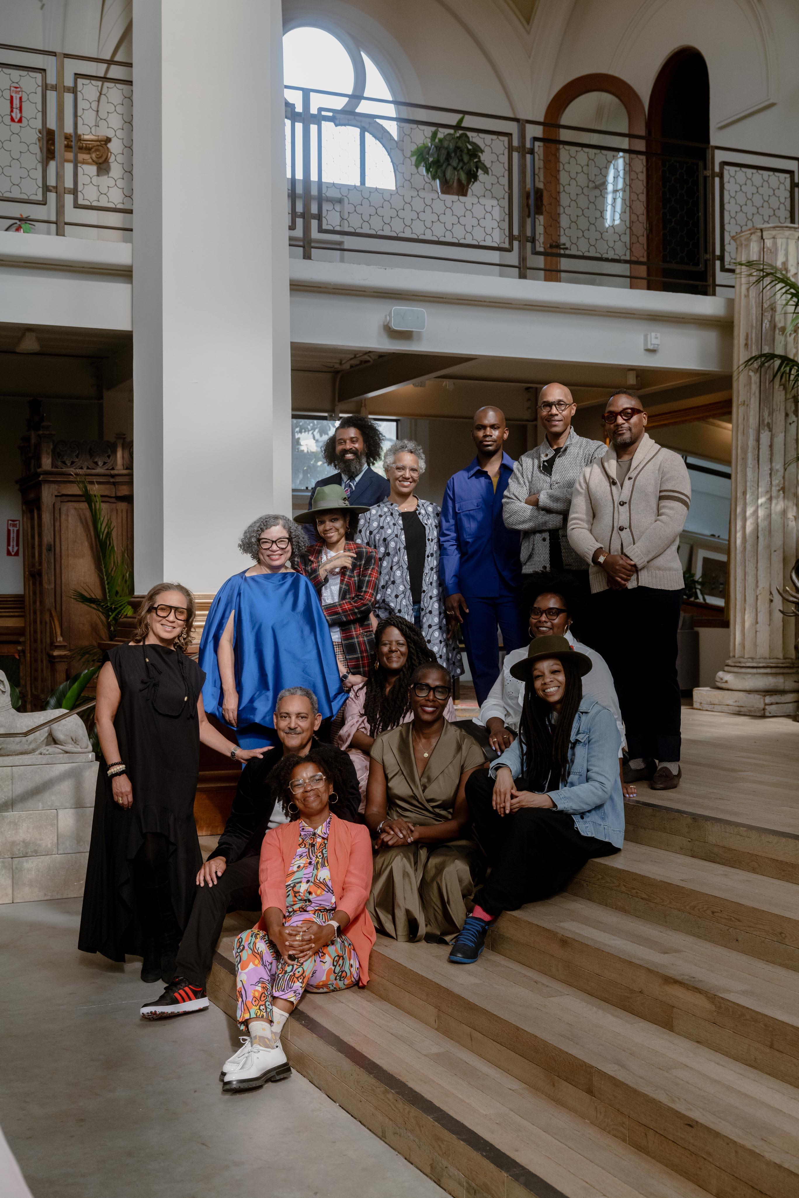 A diverse group of fourteen people poses on wooden steps in a well-lit, elegantly furnished room with large windows and archways.