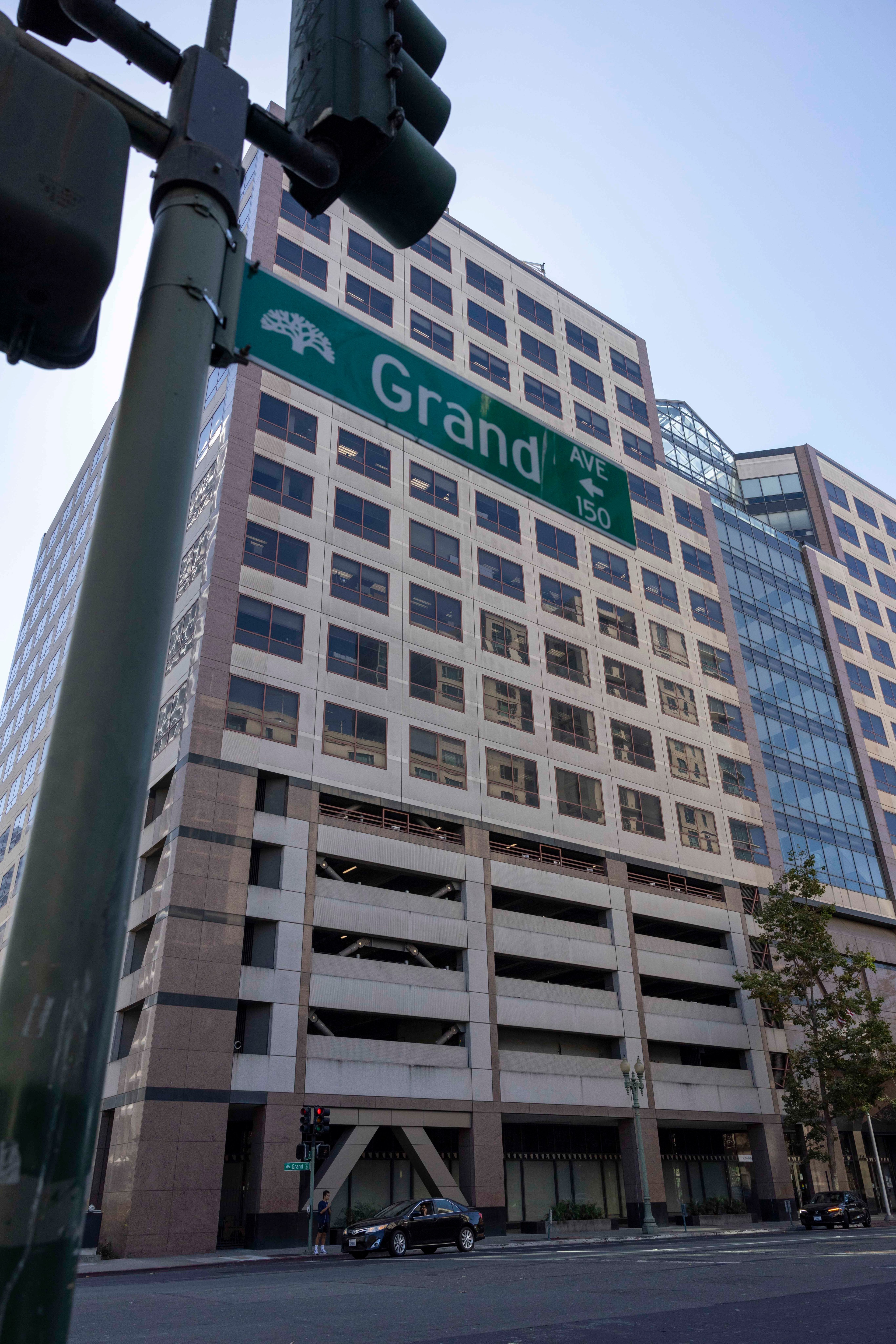 The image shows a street corner with a green &quot;Grand Ave&quot; sign attached to a traffic light pole, next to a tall office building with many windows.