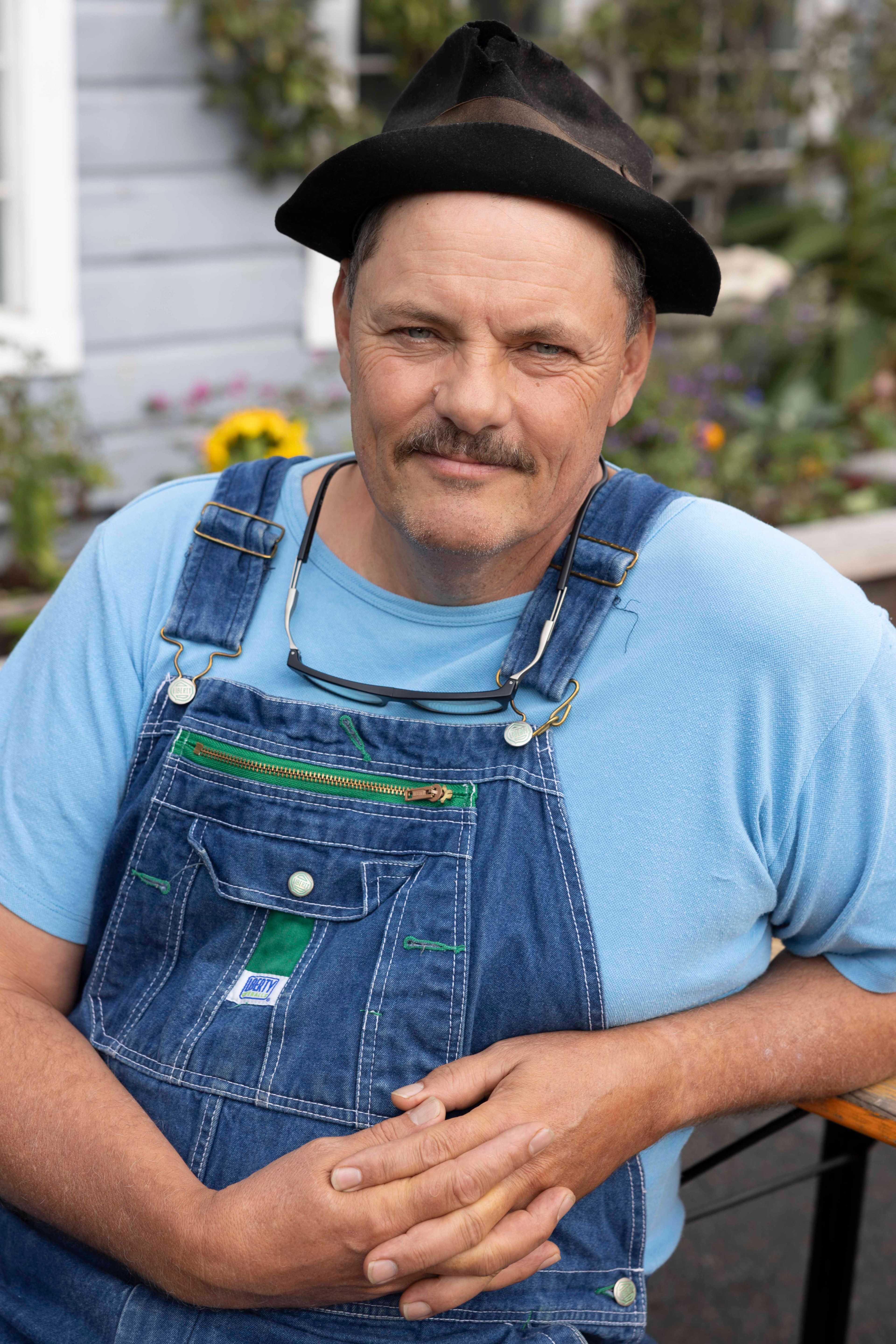 A man with a mustache wears a blue shirt, denim overalls, and a black hat, standing outside by a house with flowers and greenery in the background.