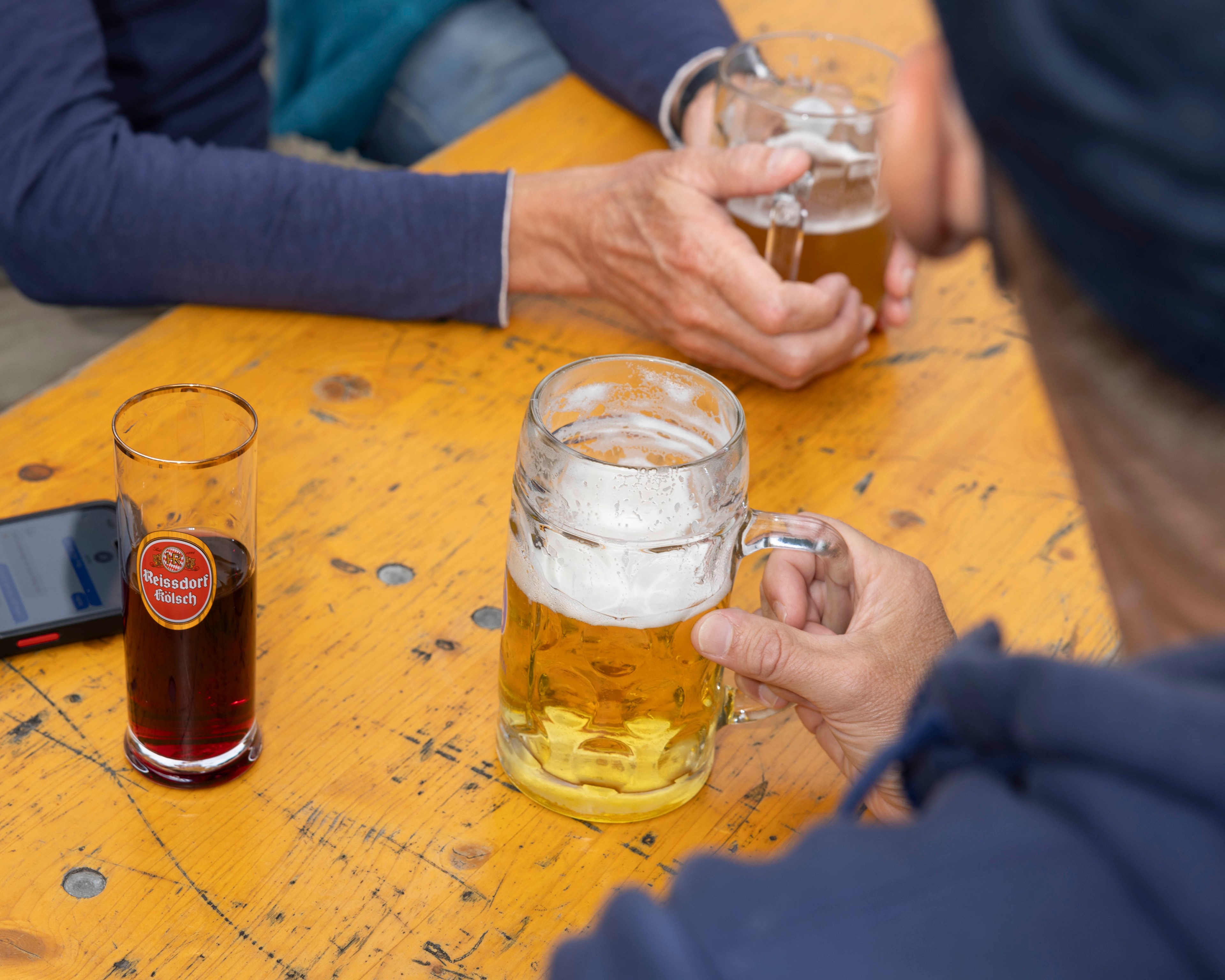 Three people are sitting around a wooden table with two holding beer mugs and one a glass of dark beverage. A smartphone lies on the table.