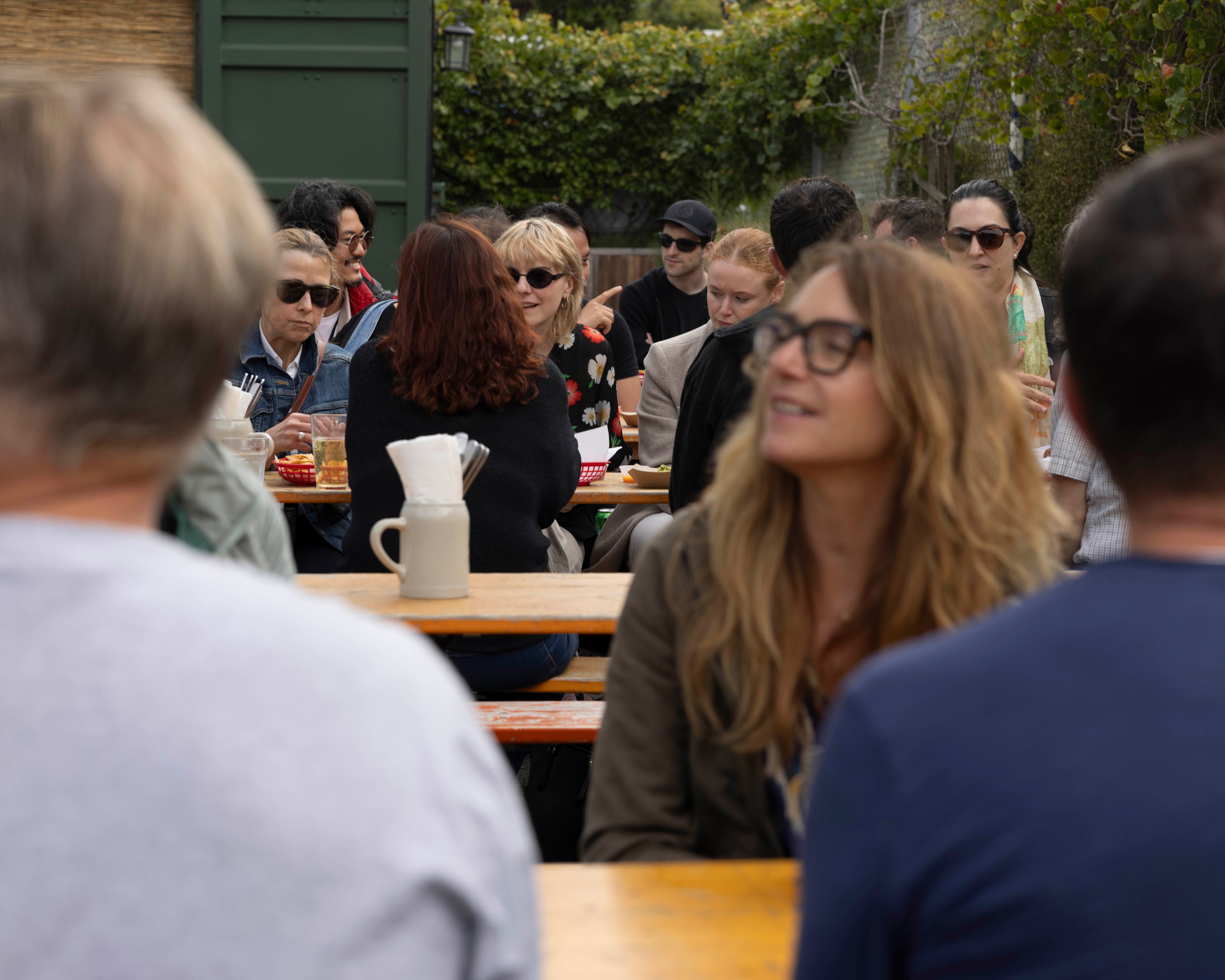 A group of people, some wearing sunglasses, are sitting at wooden picnic tables outdoors, engaged in conversation and enjoying drinks, with greenery in the background.