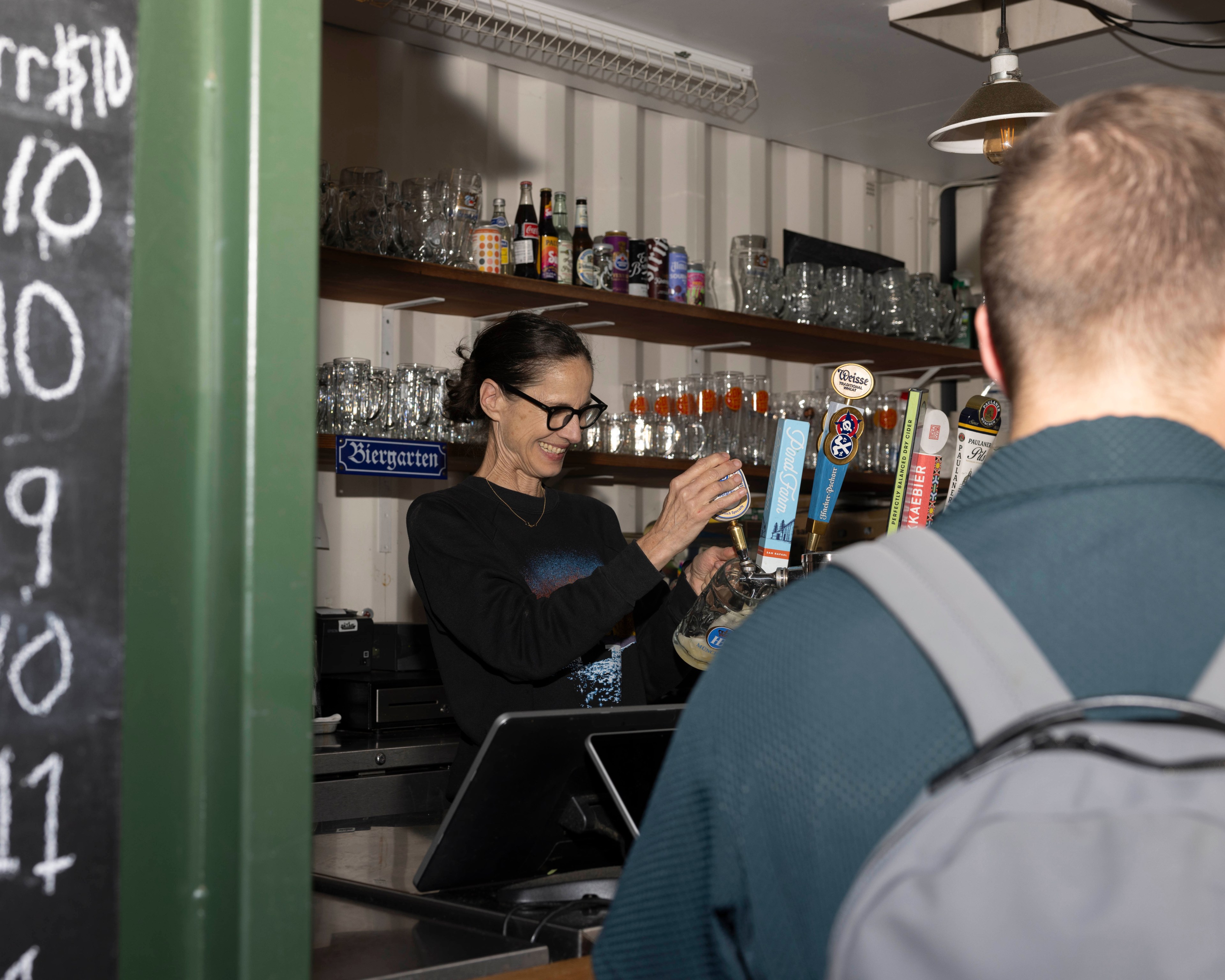 A bartender with glasses fills a glass from a beer tap, smiling. Shelves with bottles and glasses are behind her. A person with a backpack stands at the bar.