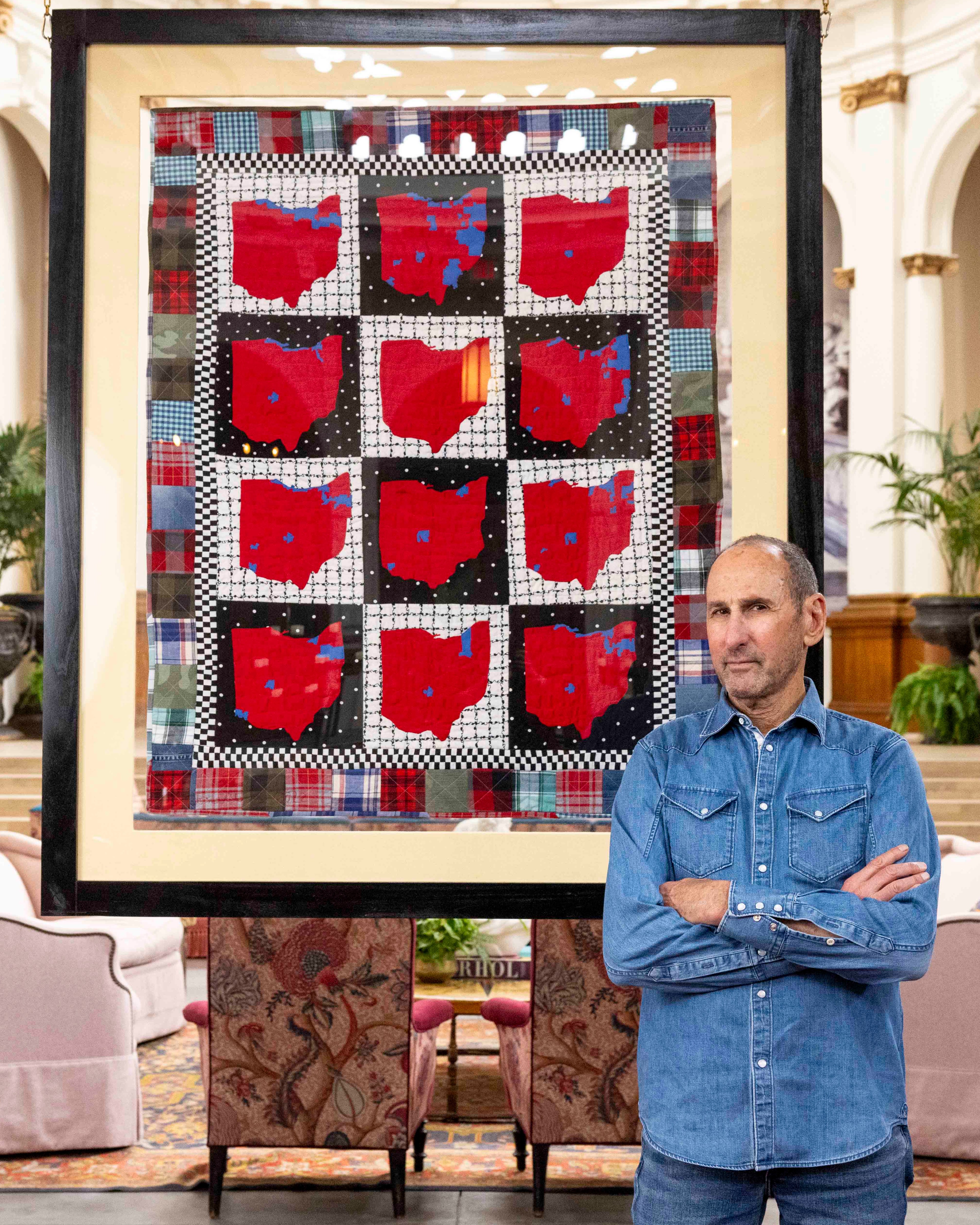 A man in a blue denim shirt stands with arms crossed next to a framed quilt with red Ohio state shapes in a grid pattern, displayed in a spacious, decorated room.