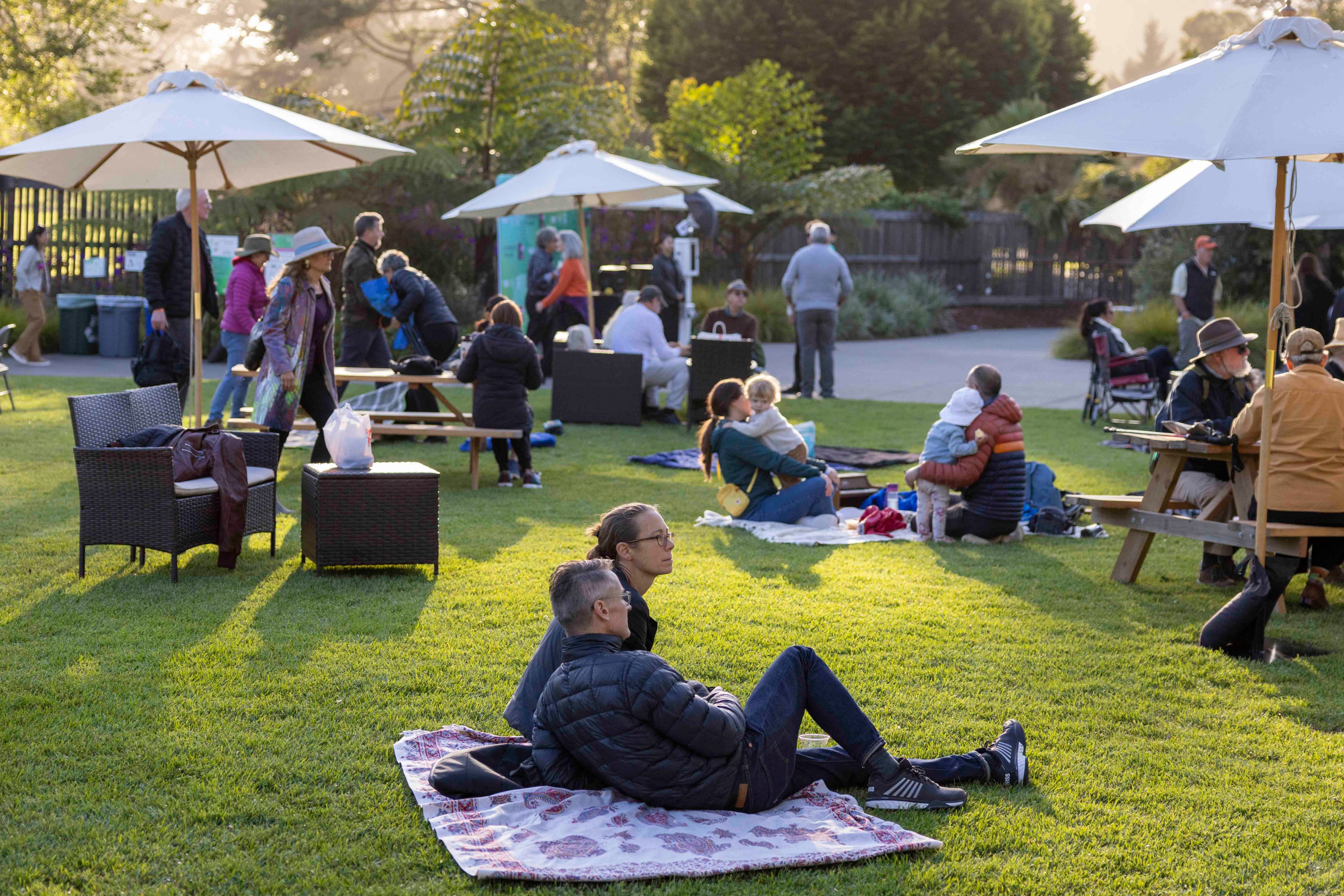 People are relaxing on a grassy area with blankets, wicker chairs, and picnic tables under sun umbrellas, enjoying a park-like setting in the late afternoon.