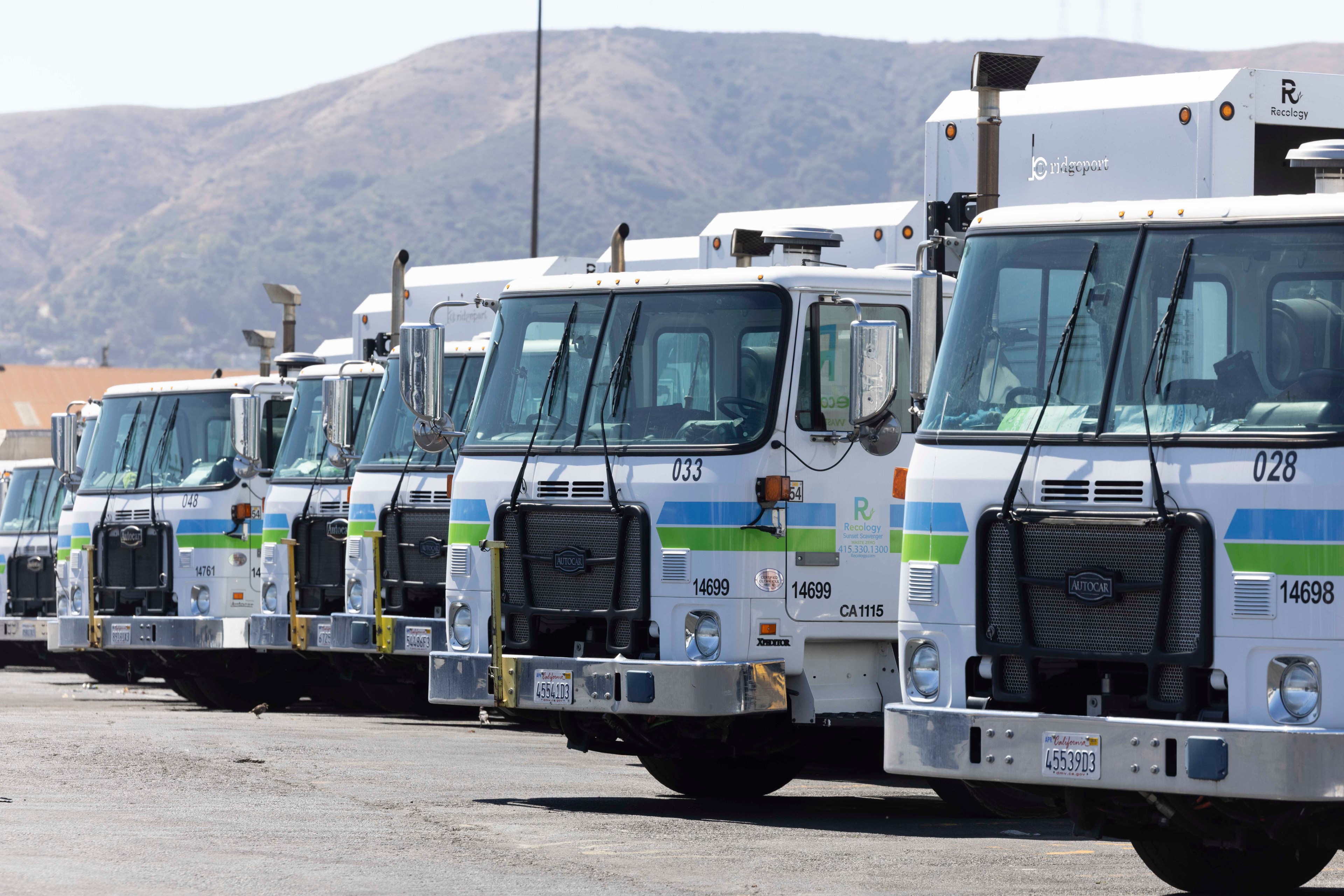 A row of white garbage trucks with green and blue stripes is parked in a lot, with hills in the background.