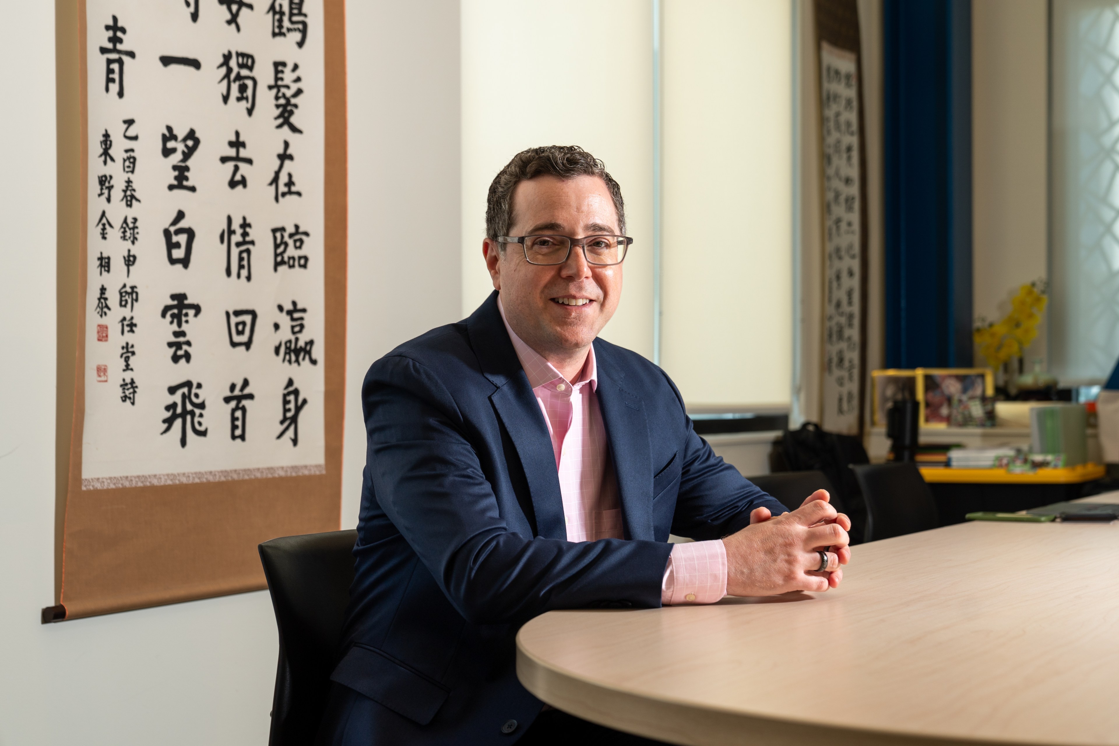 A man in a suit and glasses sits at a round table, smiling. Behind him is a wall scroll with Chinese calligraphy and an office background with books and decor.