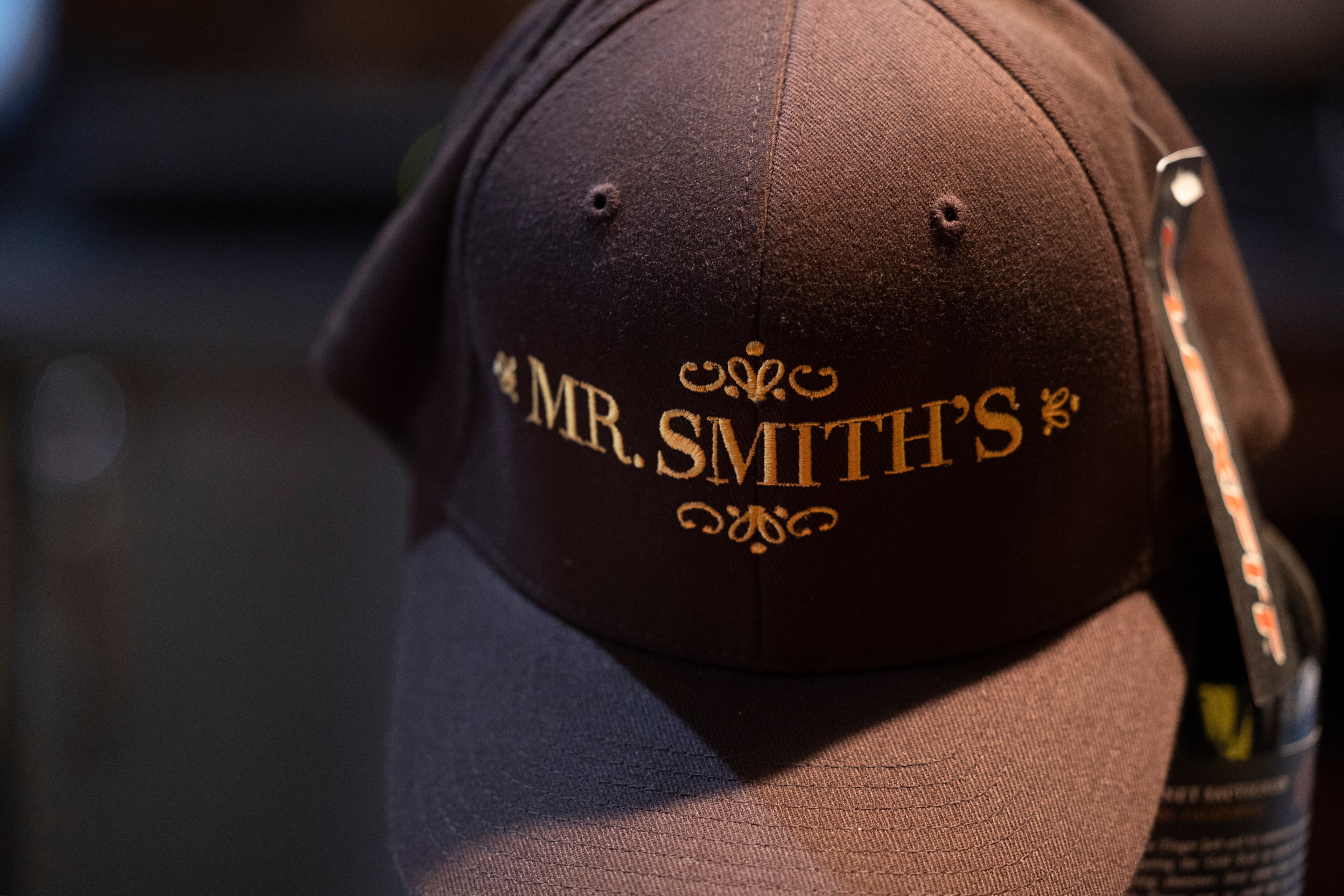 The image shows a dark brown baseball cap with the words &quot;MR. SMITH'S&quot; embroidered in gold lettering on the front.