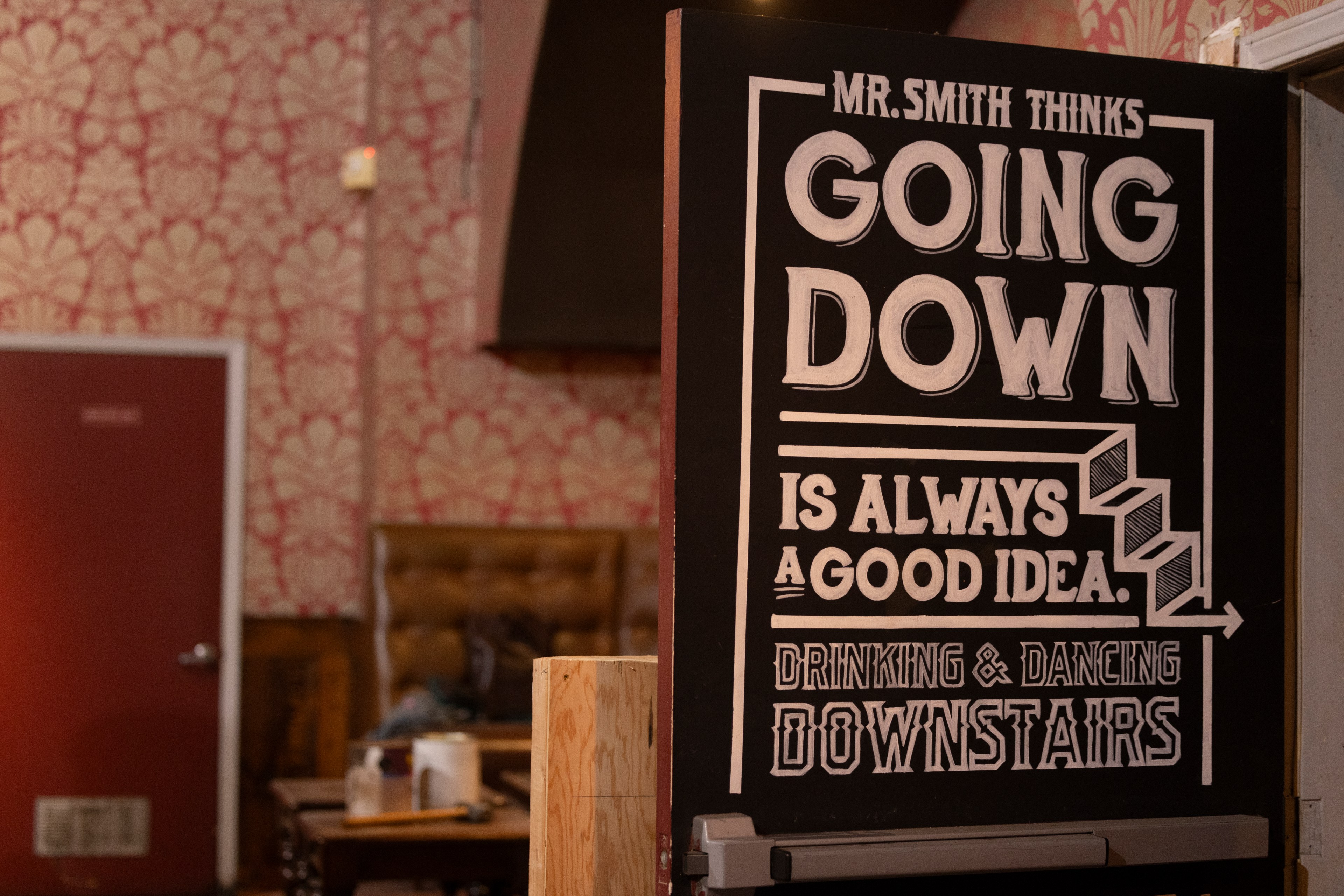 A black sign reads: &quot;Mr. Smith thinks going down is always a good idea. Drinking &amp; dancing downstairs.&quot; The background shows patterned wallpaper and a closed red door.