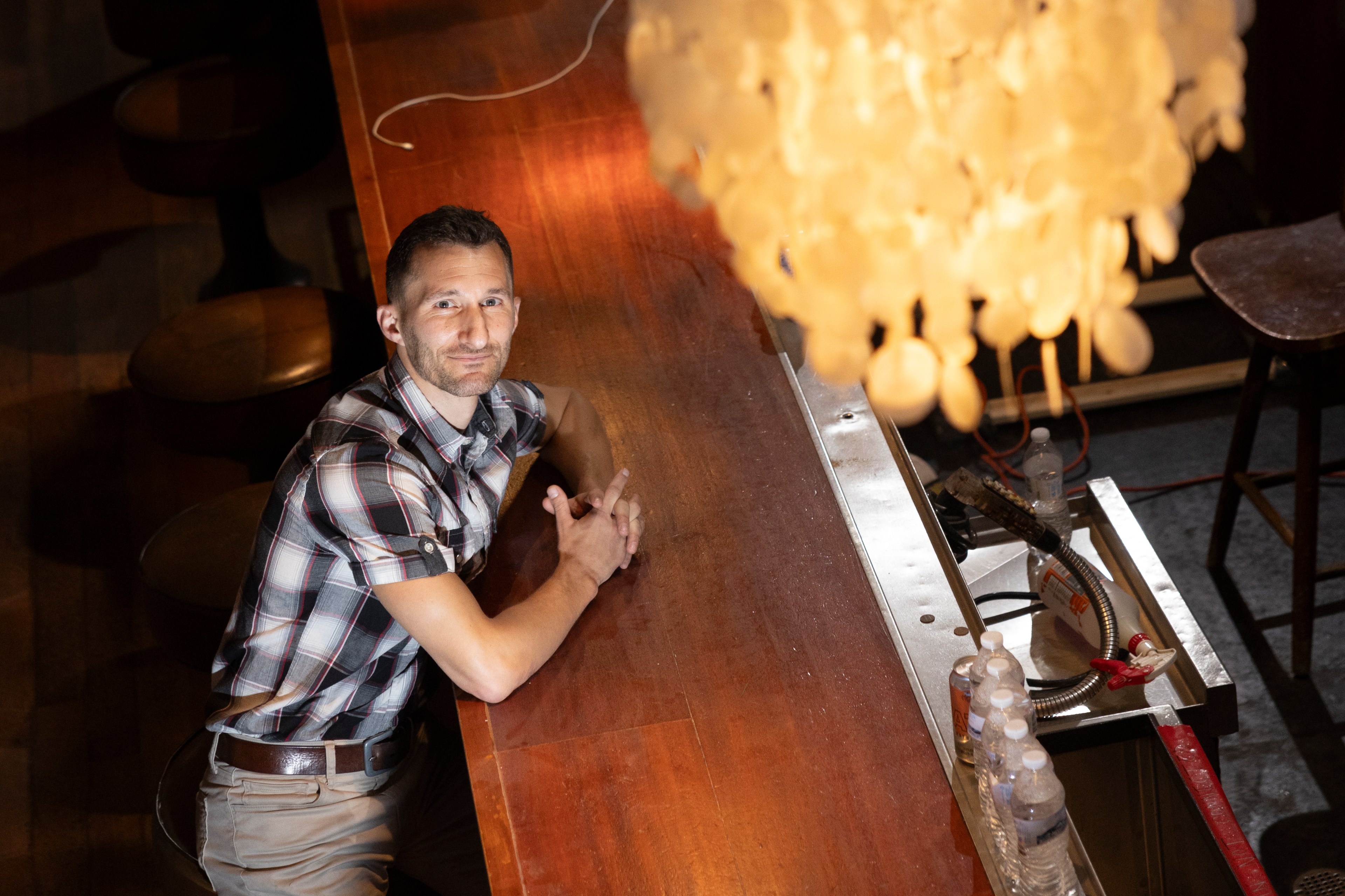 A man in a plaid shirt leans against a bar counter, illuminated by large, ornate chandeliers hanging overhead. The setting is cozy with a warm ambiance.