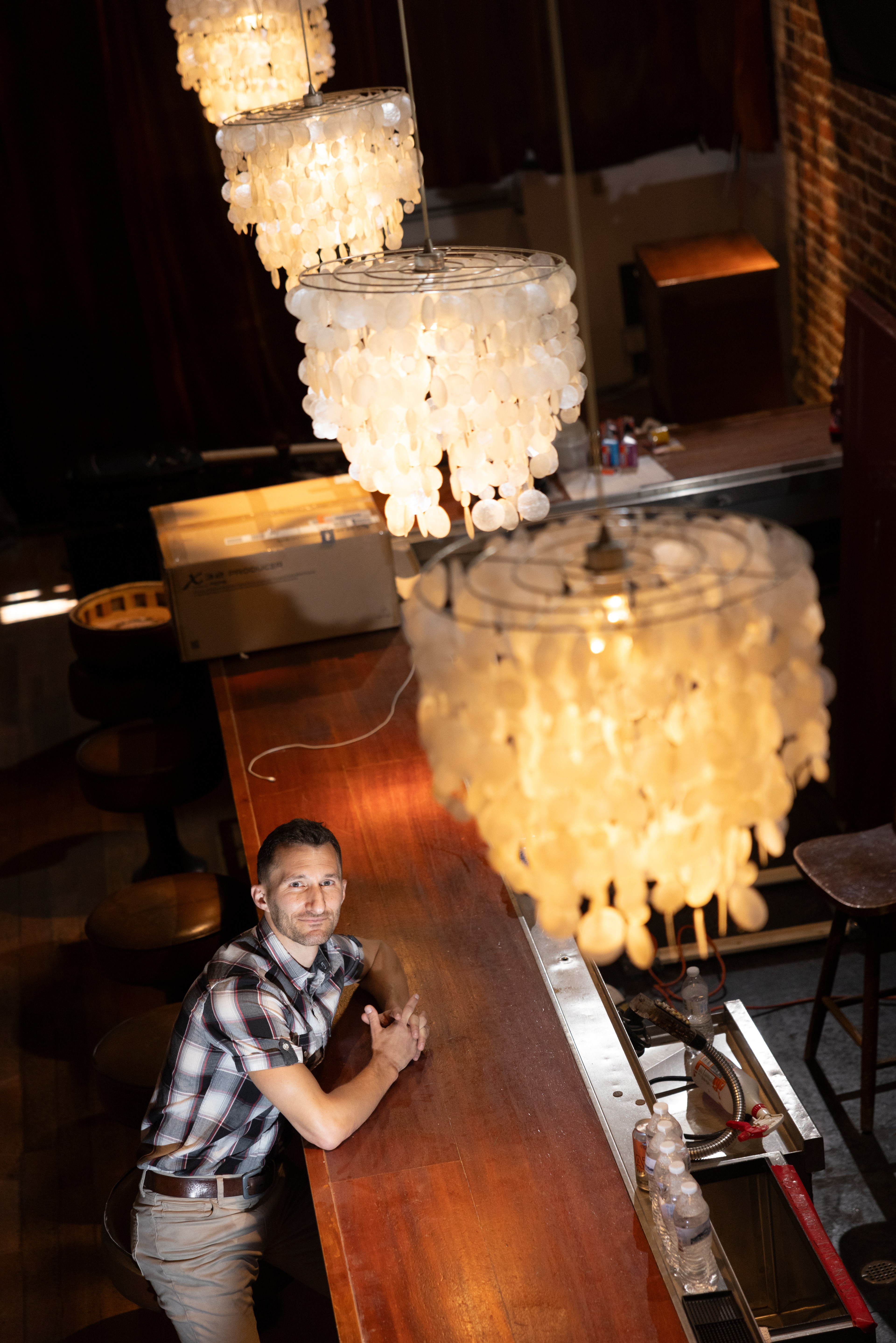 A man in a plaid shirt leans against a bar counter, illuminated by large, ornate chandeliers hanging overhead. The setting is cozy with a warm ambiance.