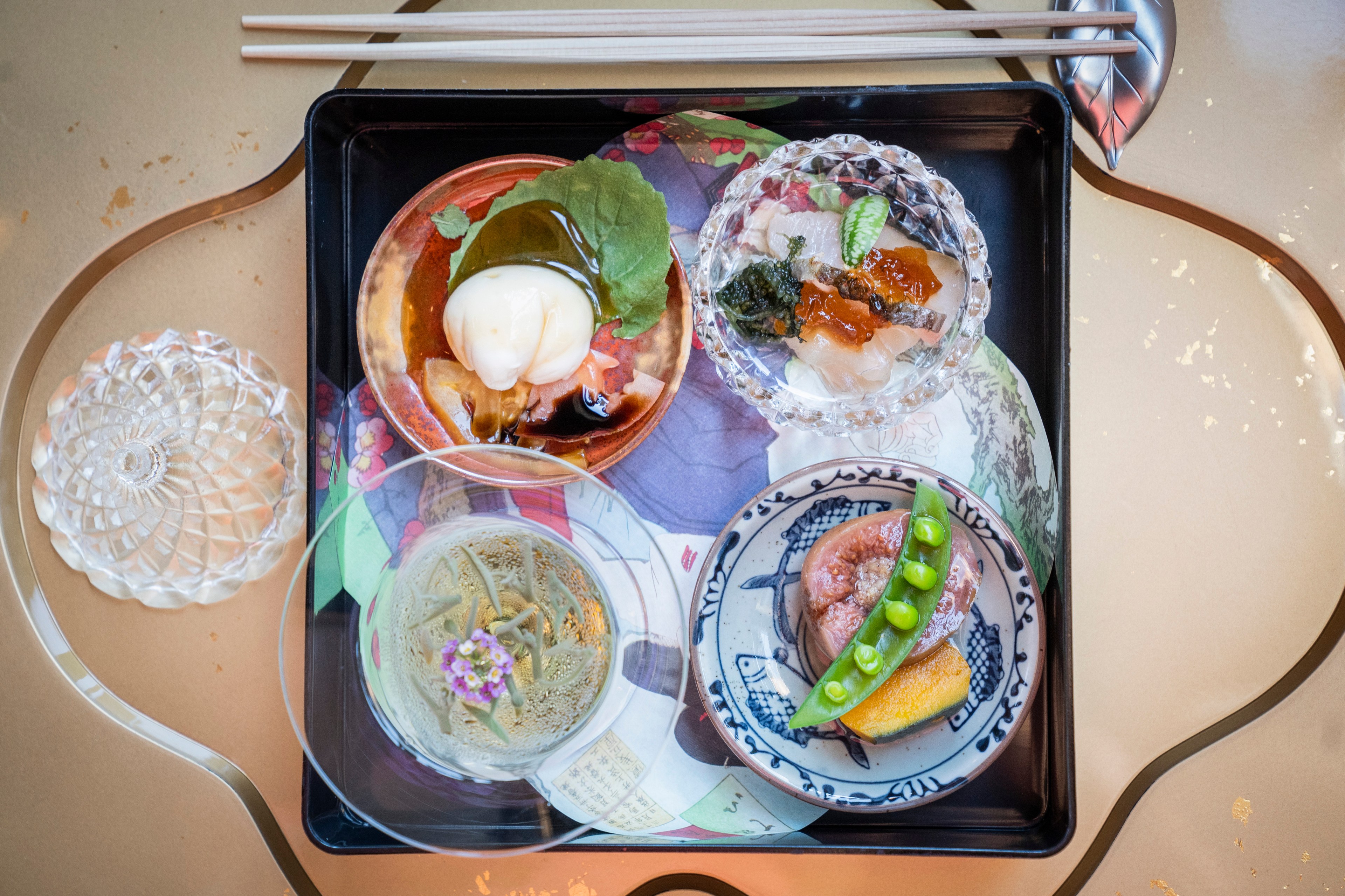 A tray with four elegant bowls containing various foods, chopsticks, and a glass dish, set on a reflective, gold-patterned table.