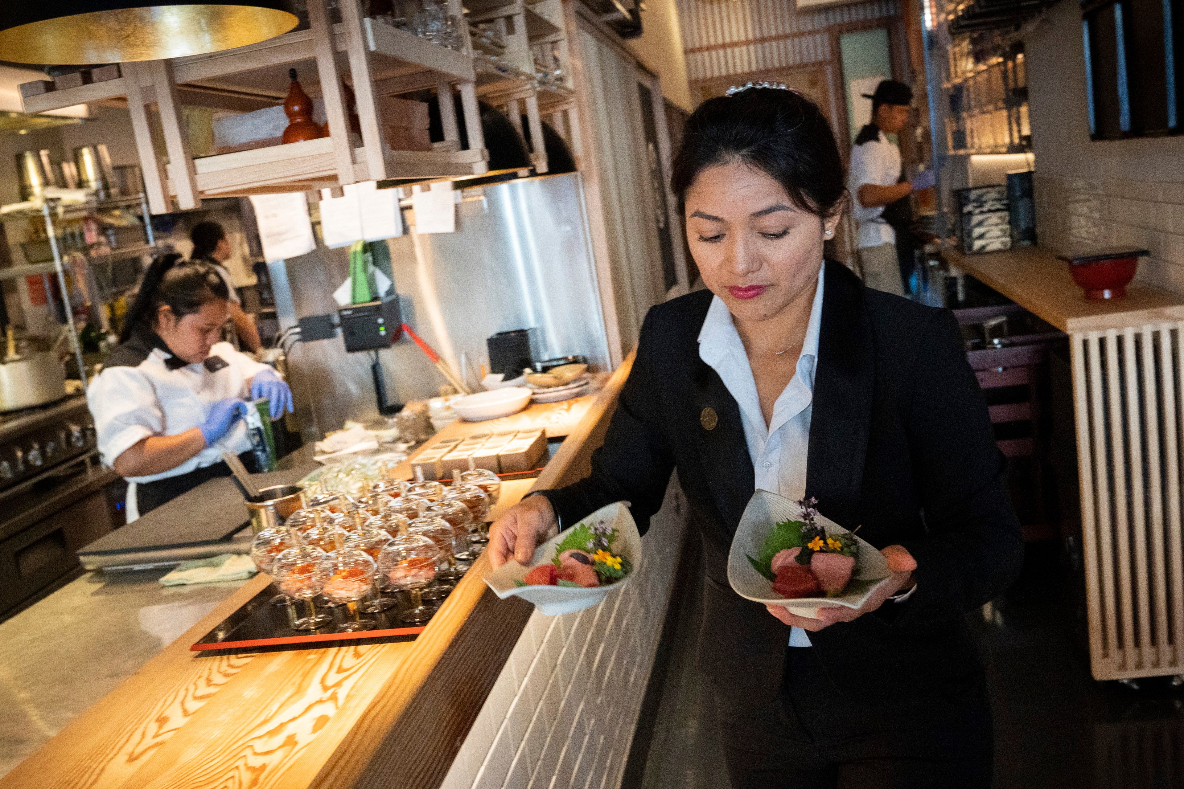 A server in a black suit and white shirt carries two dishes with garnished food in a restaurant; other staff work in the background near a counter with more dishes.