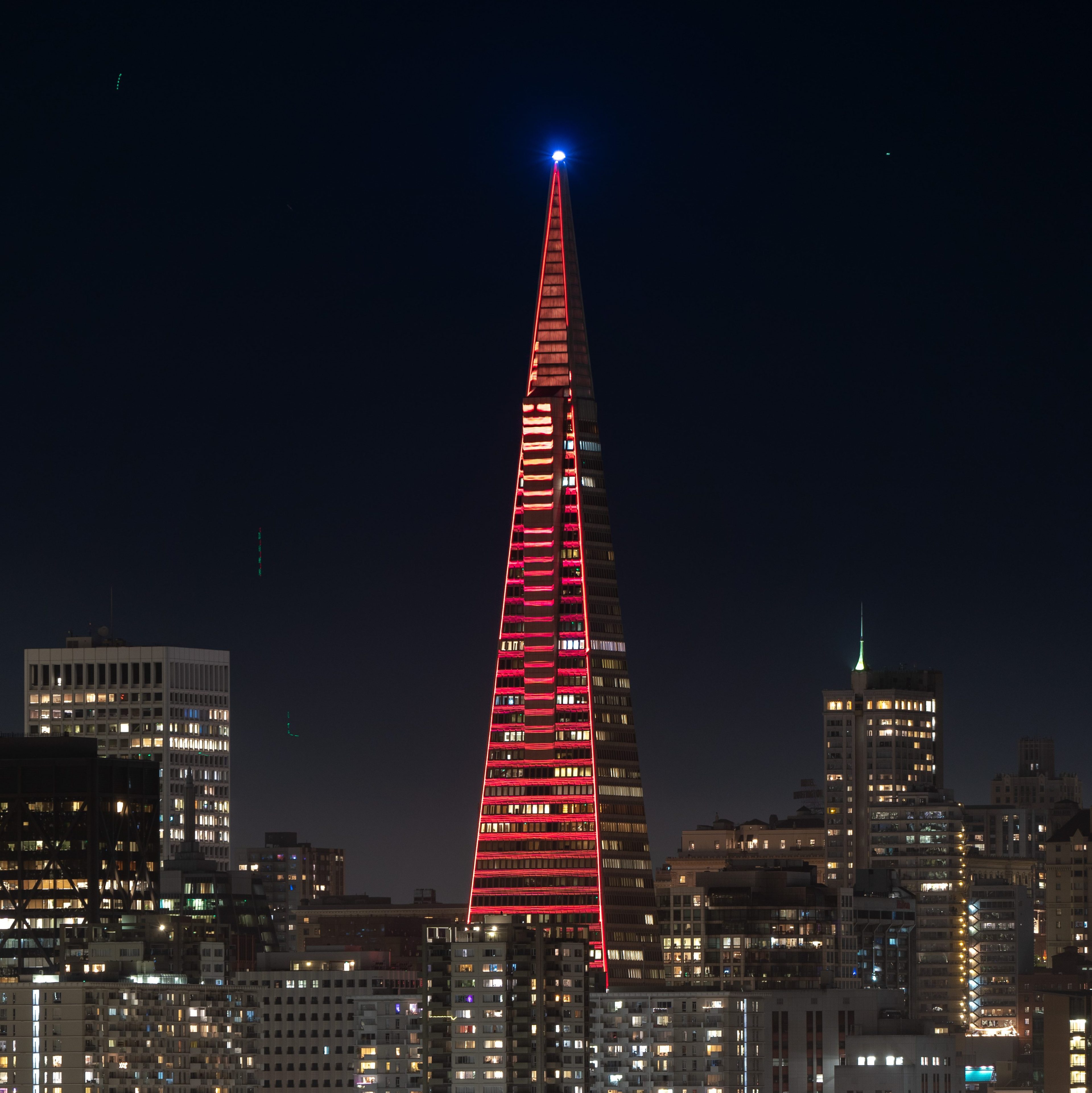 A tall, triangular building with red and white lights stands prominently at night, surrounded by other high-rise buildings with illuminated windows.