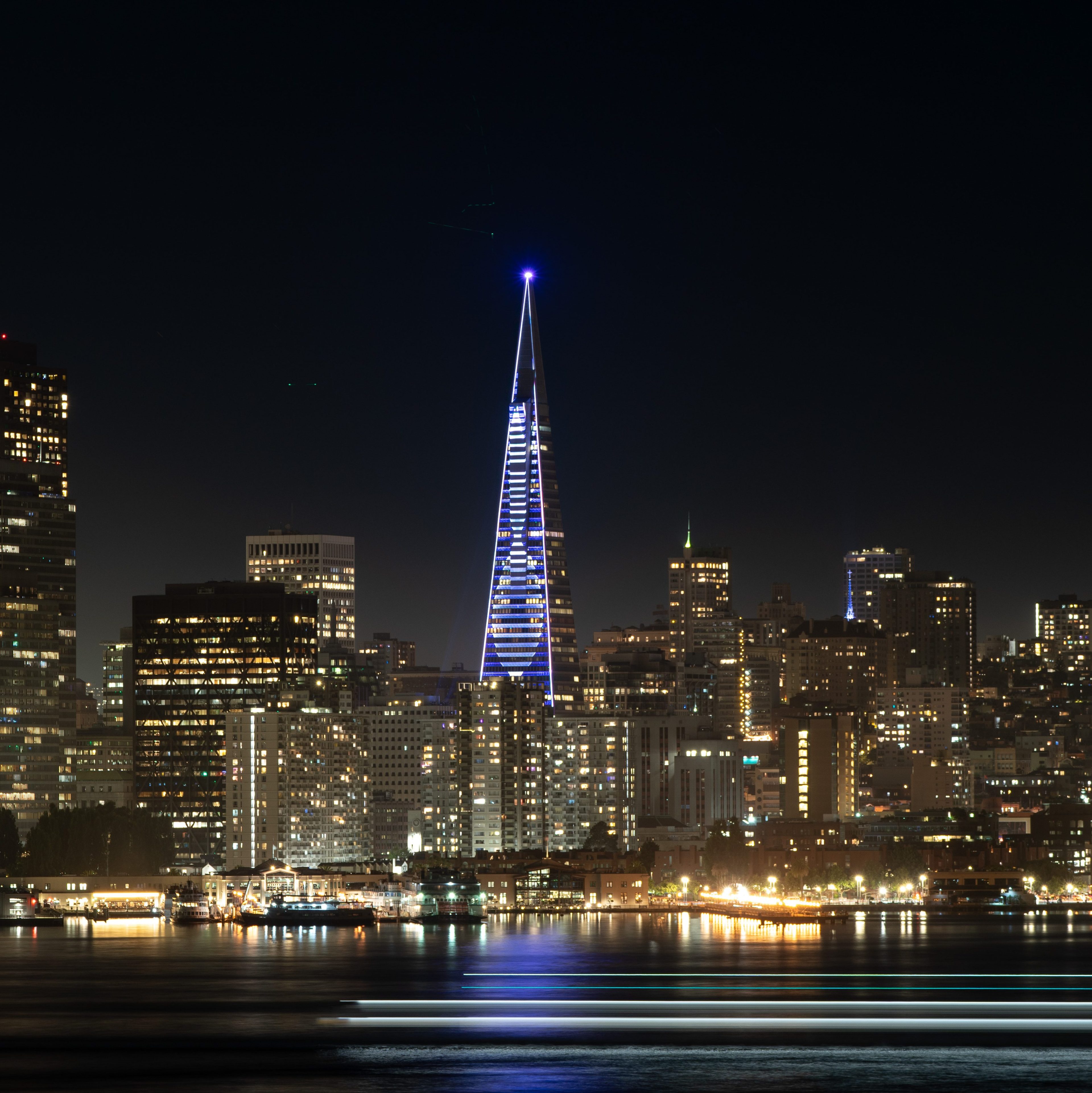 The image shows a city skyline at night with brightly lit buildings, dominated by a tall pyramid-shaped skyscraper illuminated in blue lights.