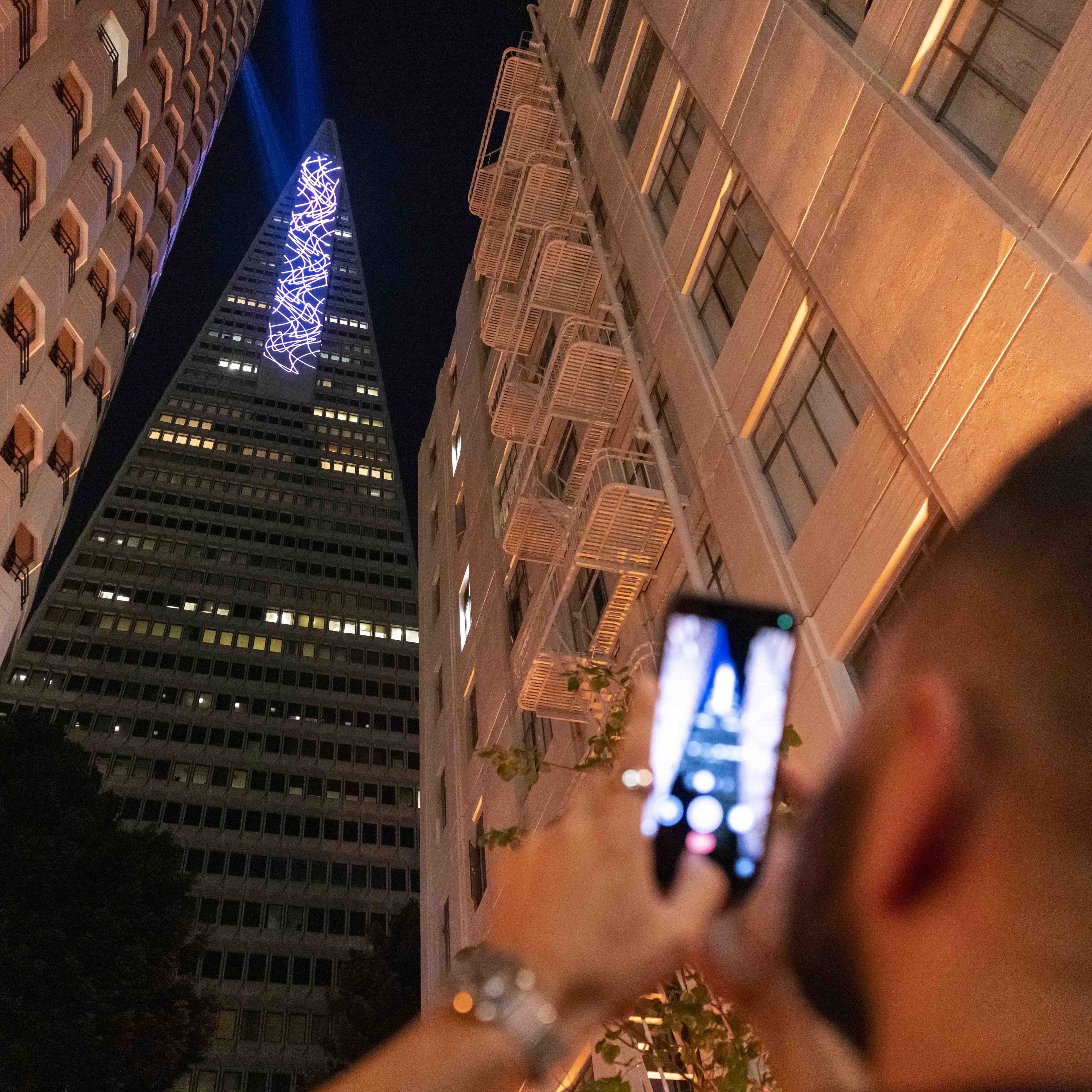 A person is photographing a tall, triangular skyscraper adorned with blue lights at night, framed by surrounding buildings with visible fire escapes.