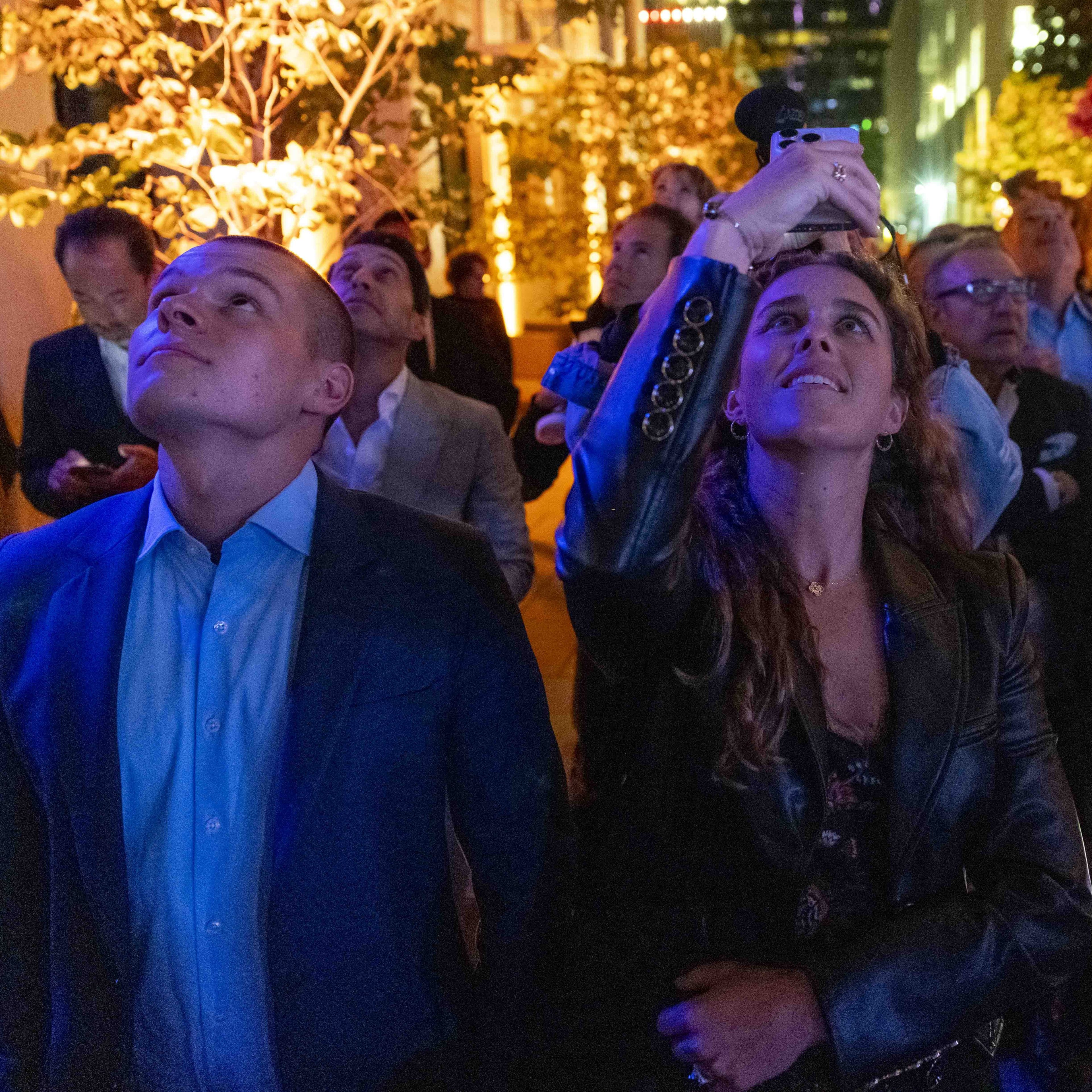 A group of people are looking upwards at night, with some holding their phones up. They are outdoors with illuminated trees and city lights in the background.