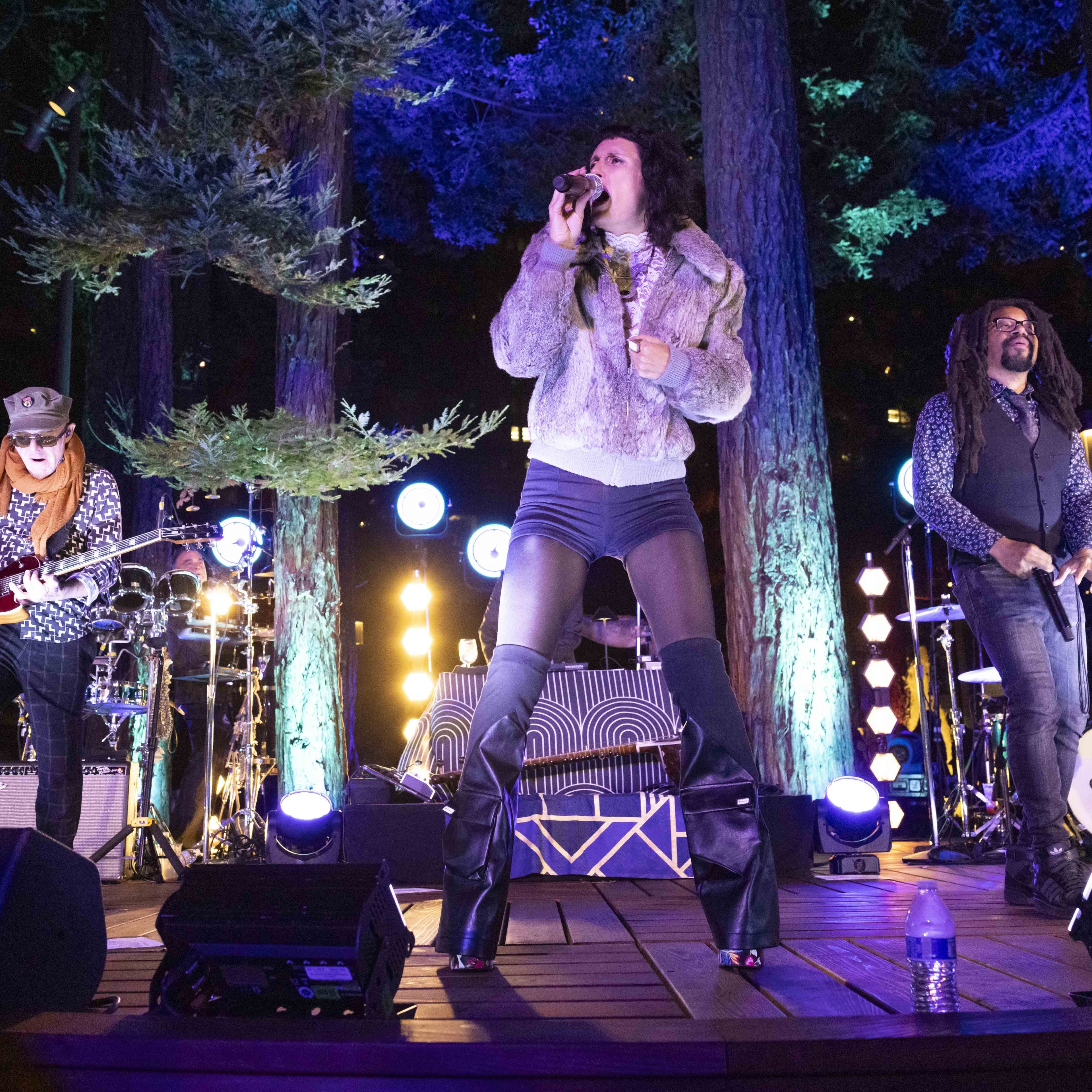 Three musicians perform on an outdoor, forest-lit stage; a guitarist on the left, a singer in the center with a mic, and another vocalist on the right.