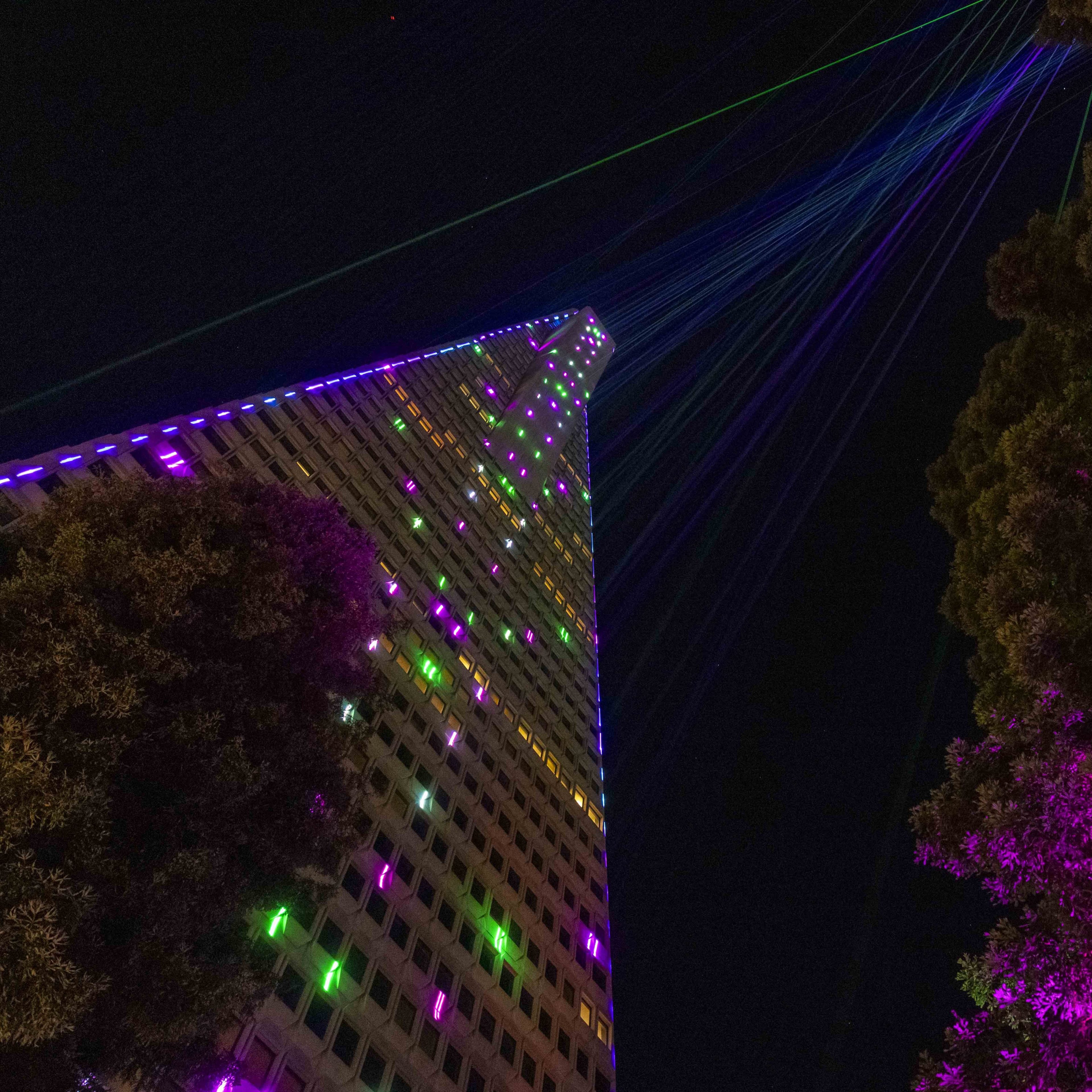 A tall building is brightly illuminated with multi-colored lights and lasers, set against a dark night sky, with trees framing the sides.