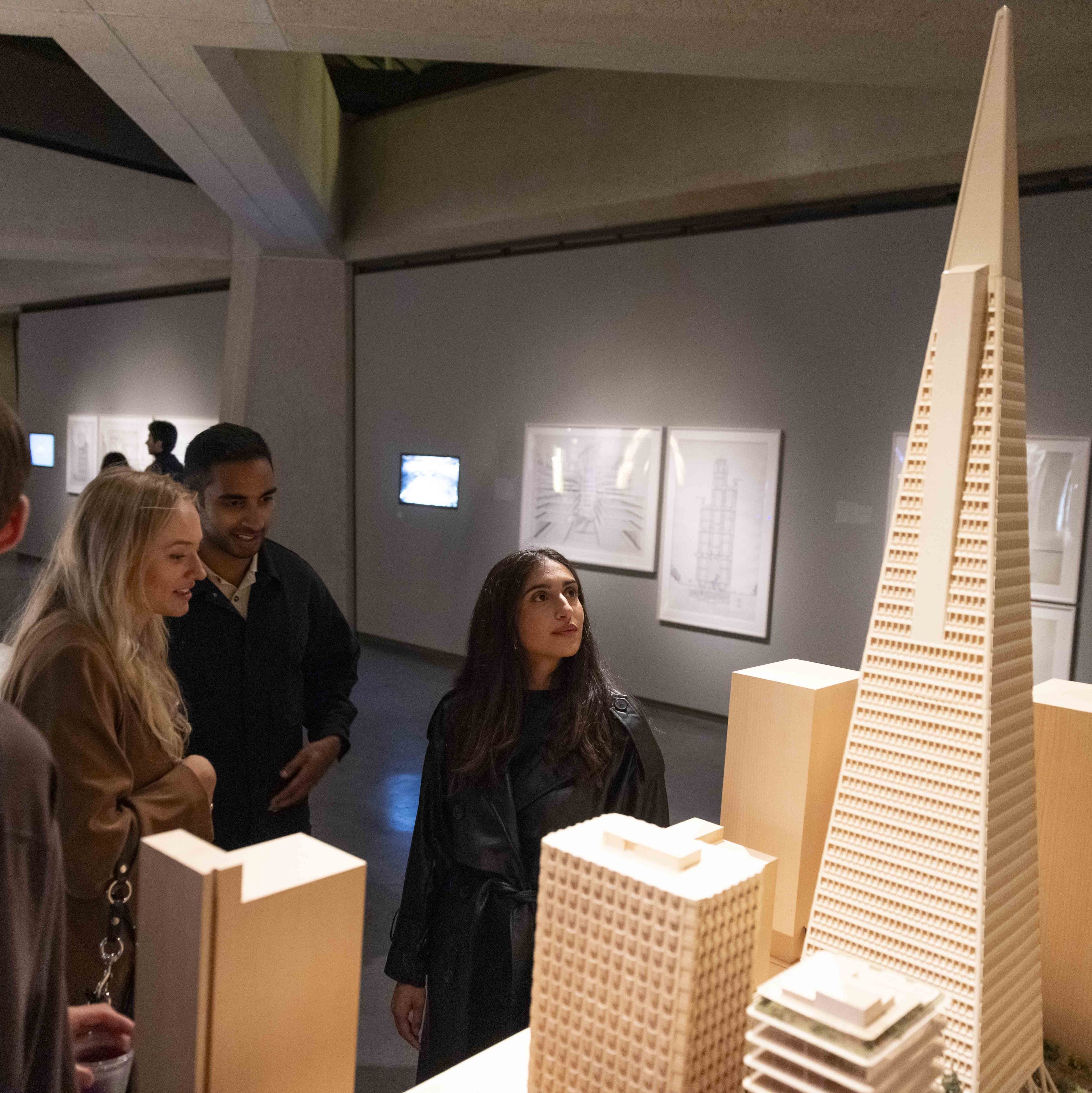 Three people admire a detailed architectural model of tall buildings in a dimly lit museum, with framed sketches and a small screen displayed on the walls behind them.