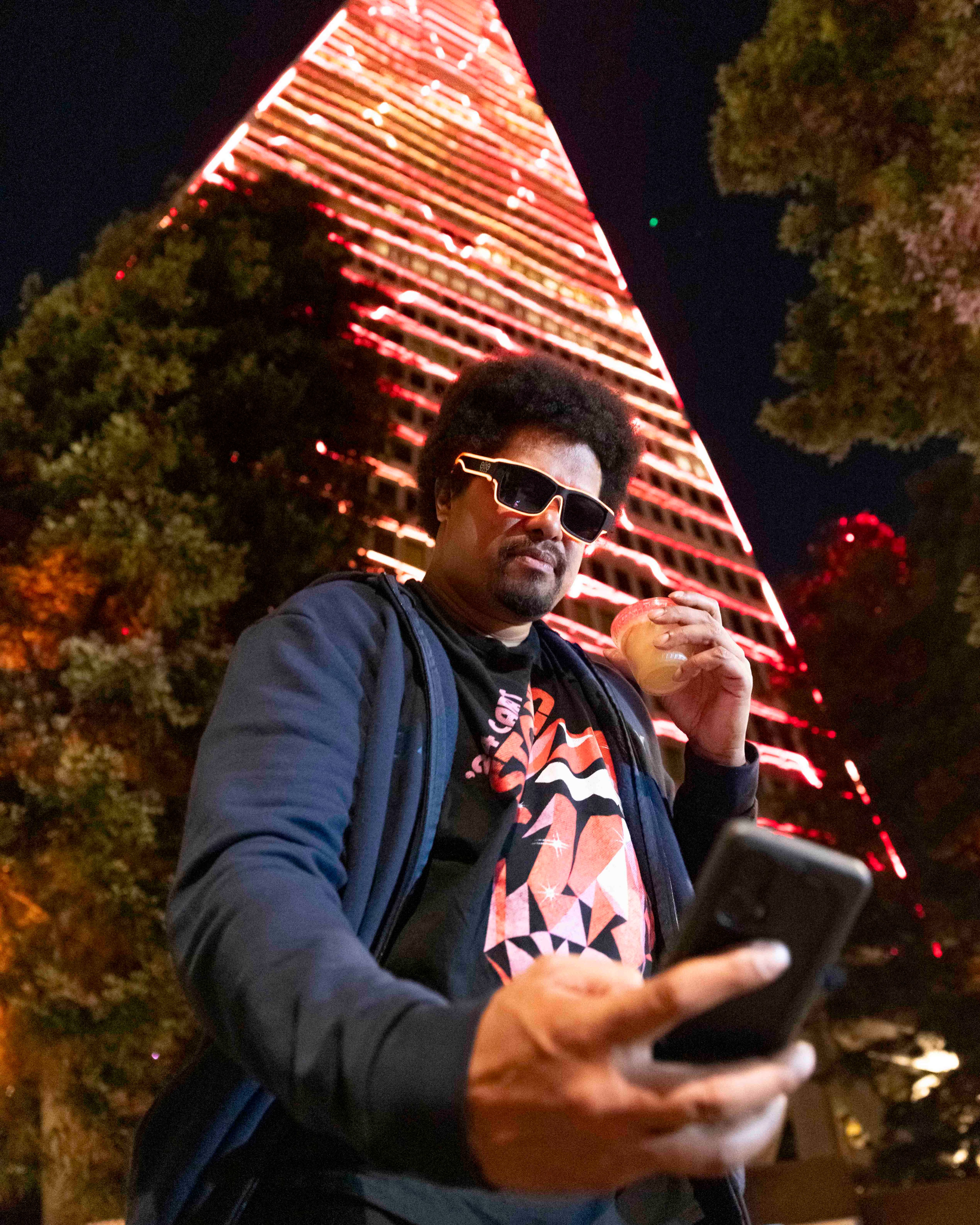 A man wearing sunglasses holds a drink in one hand and a phone in the other, posing in front of a tall, illuminated, triangular building at night.