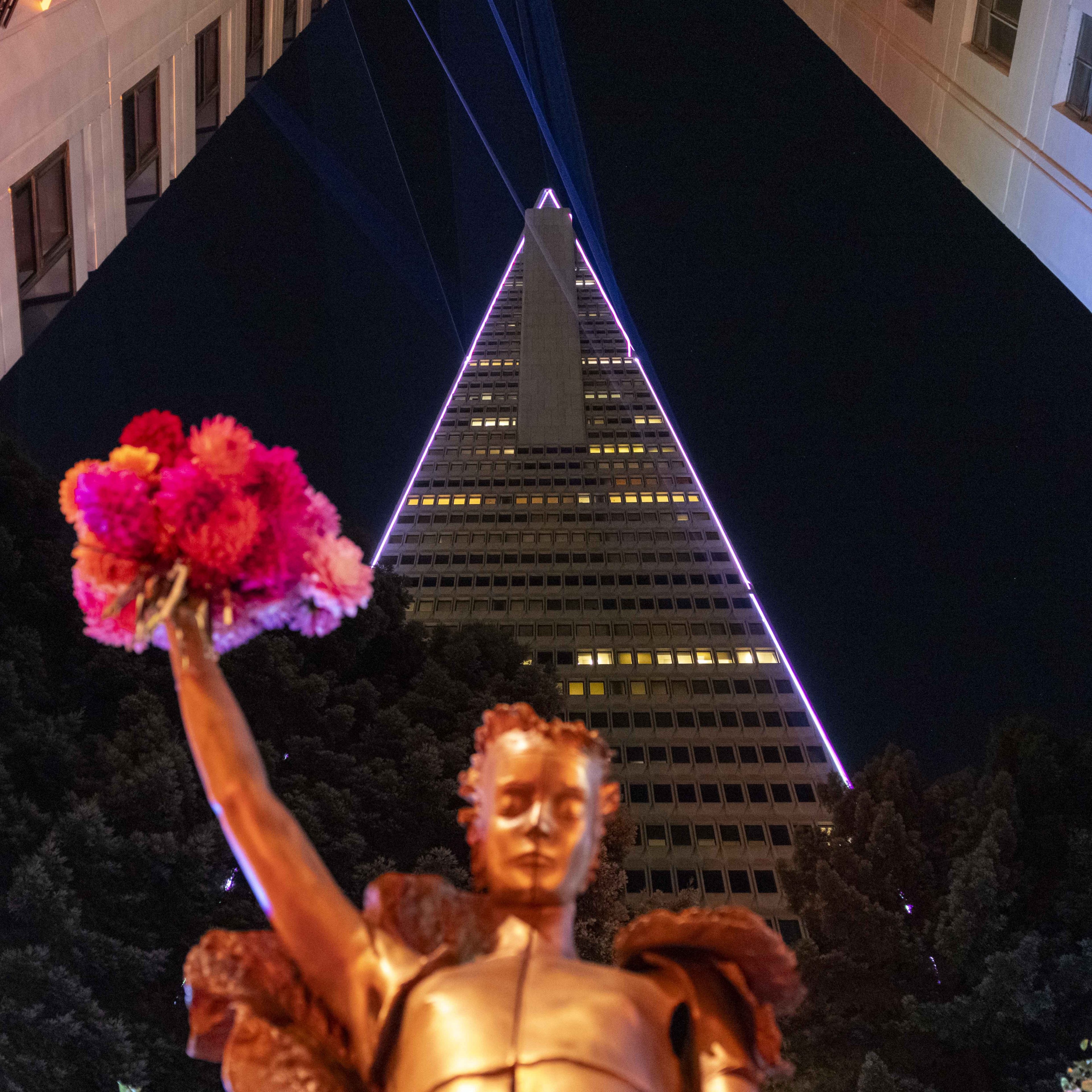 A bronze-colored statue holds a bouquet of vivid pink and red flowers in front of a tall, illuminated triangular skyscraper under a dark night sky.