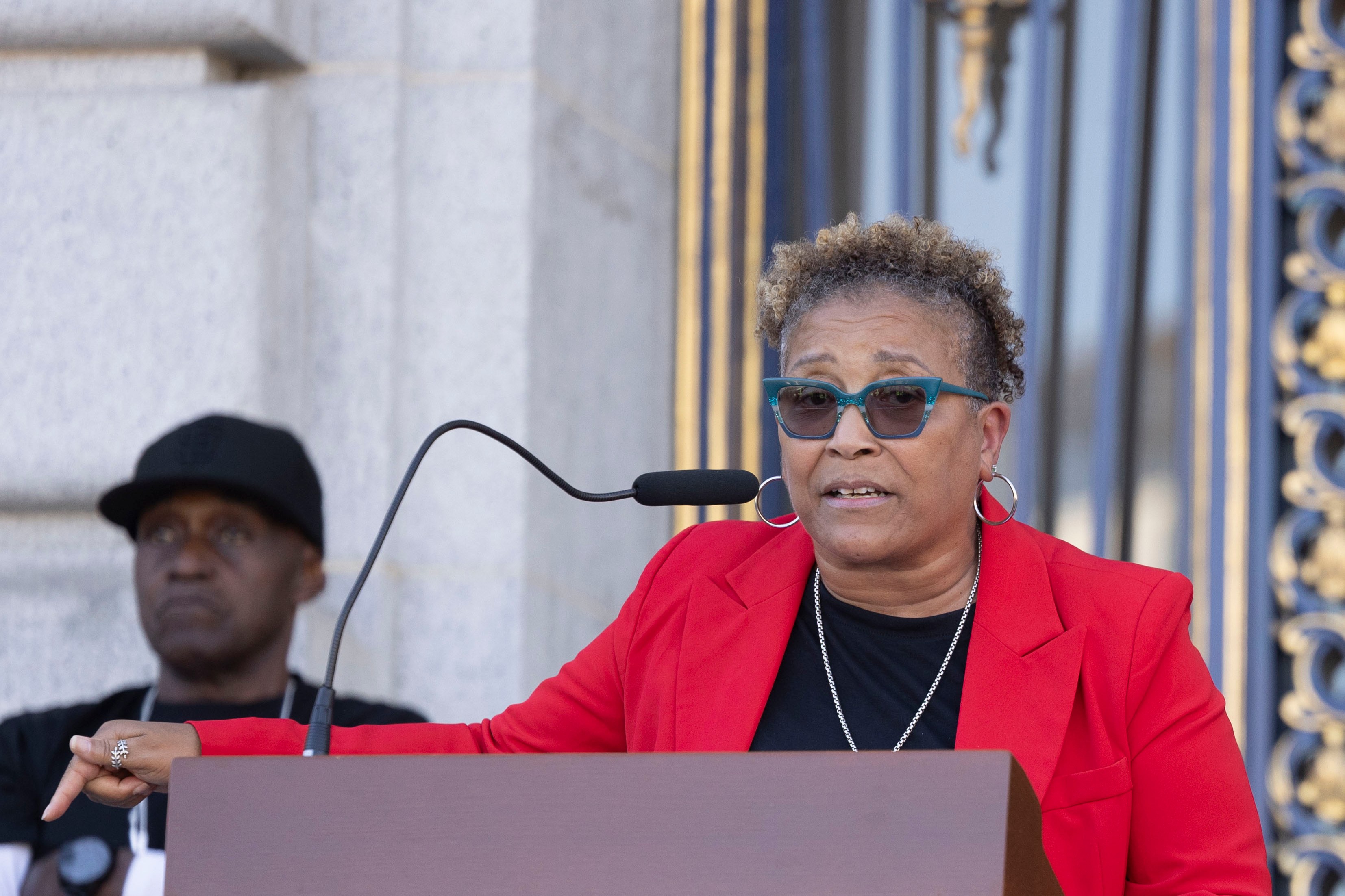 A person in a red blazer speaks at a podium with a microphone. Another individual stands in the background, dressed in black with a cap.