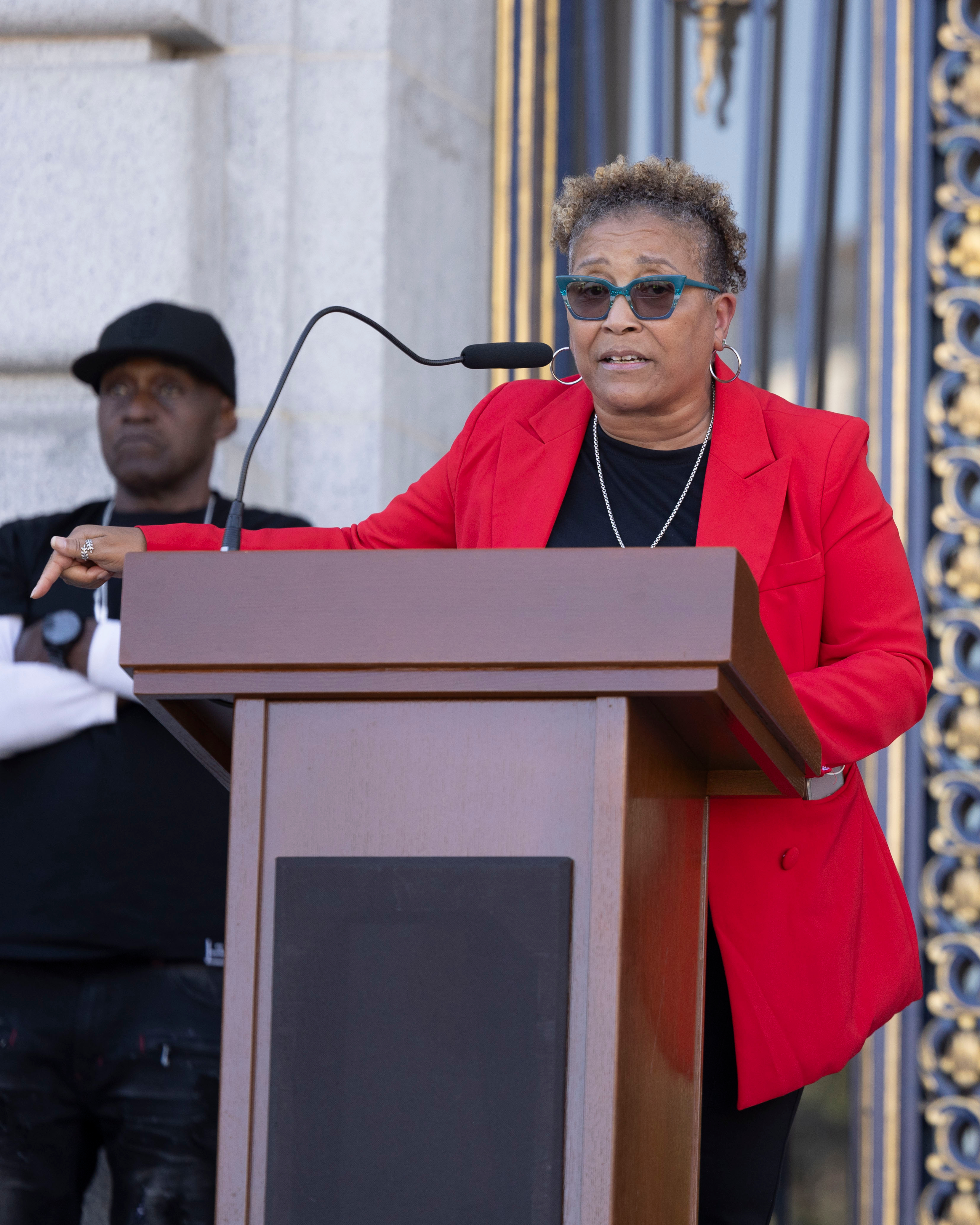 A person in a red blazer speaks at a podium with a microphone. Another individual stands in the background, dressed in black with a cap.