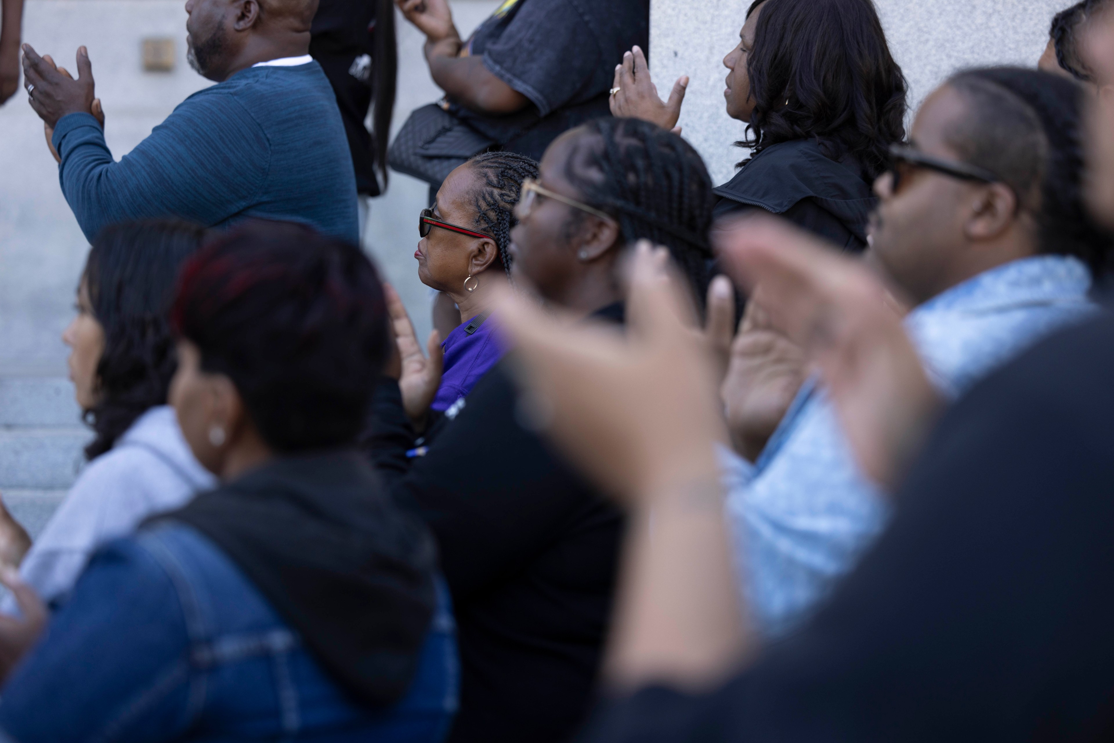 A group of people attentively listens and claps in an outdoor setting, some wearing glasses and varying in attire such as jackets and sweaters.