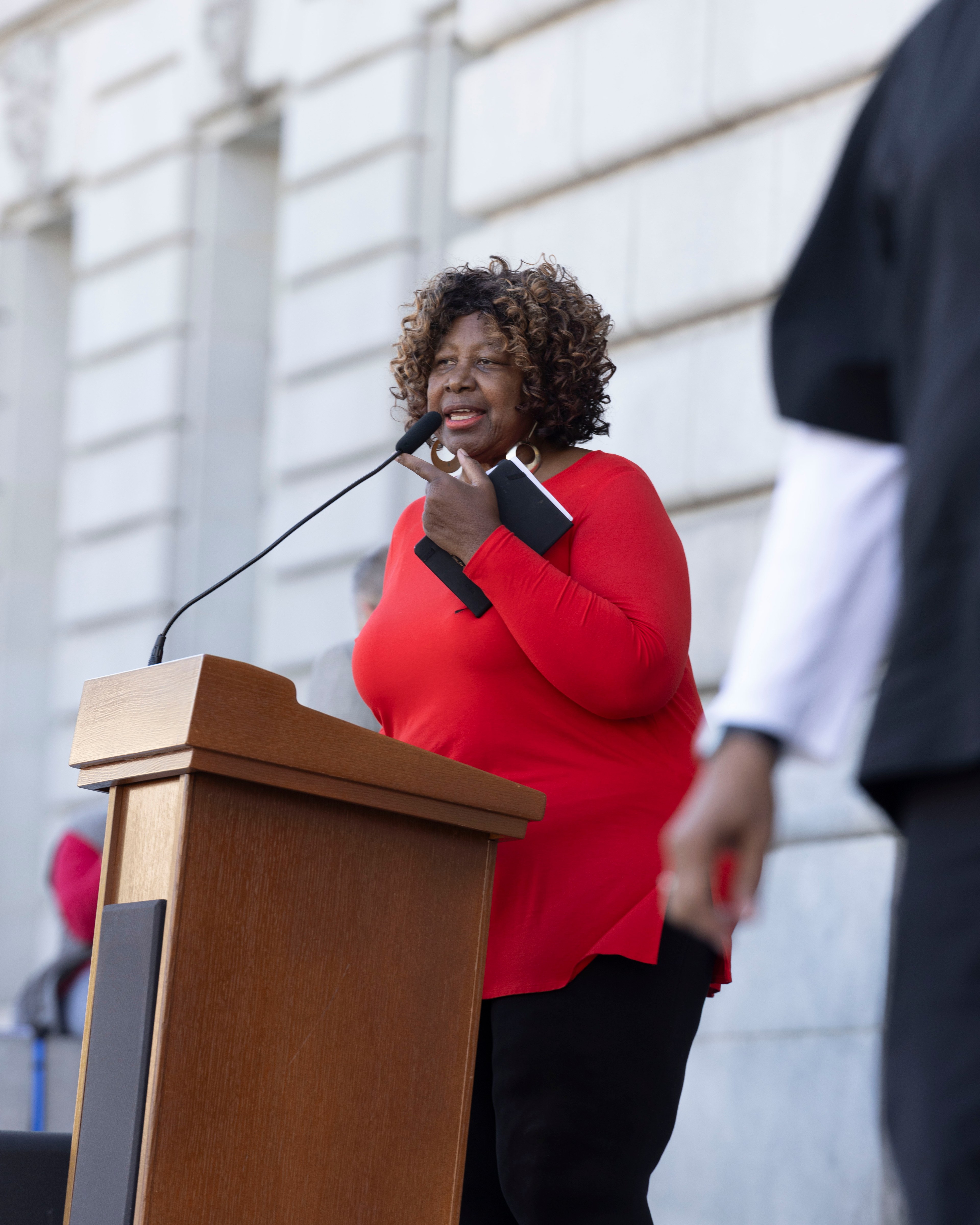 A woman in a red top speaks at a podium outdoors, holding a booklet. She stands in front of a large stone building, and another person is partially visible nearby.