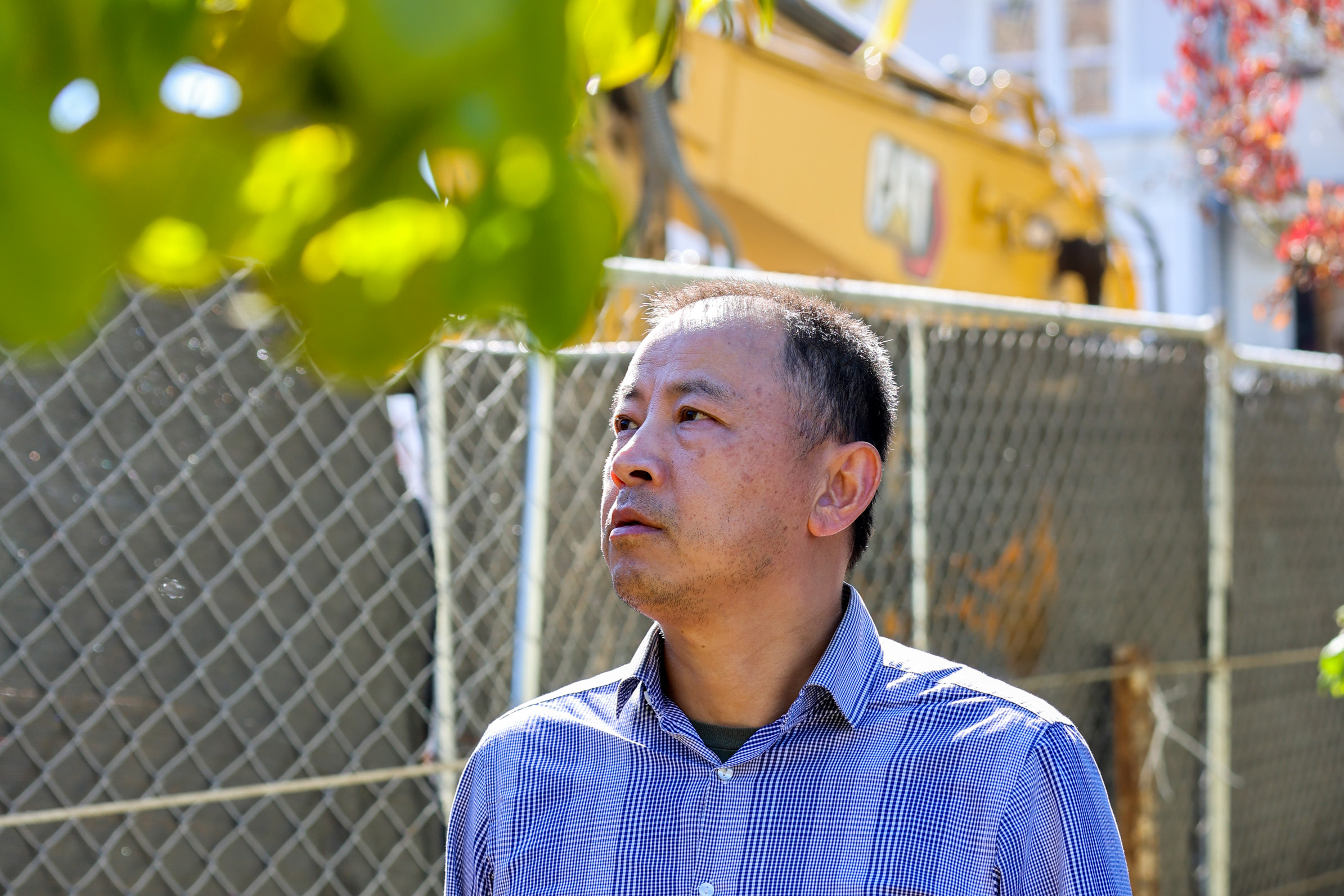 A man in a checkered shirt looks thoughtful, standing outdoors by a chain-link fence, with construction equipment and blurred foliage in the background.