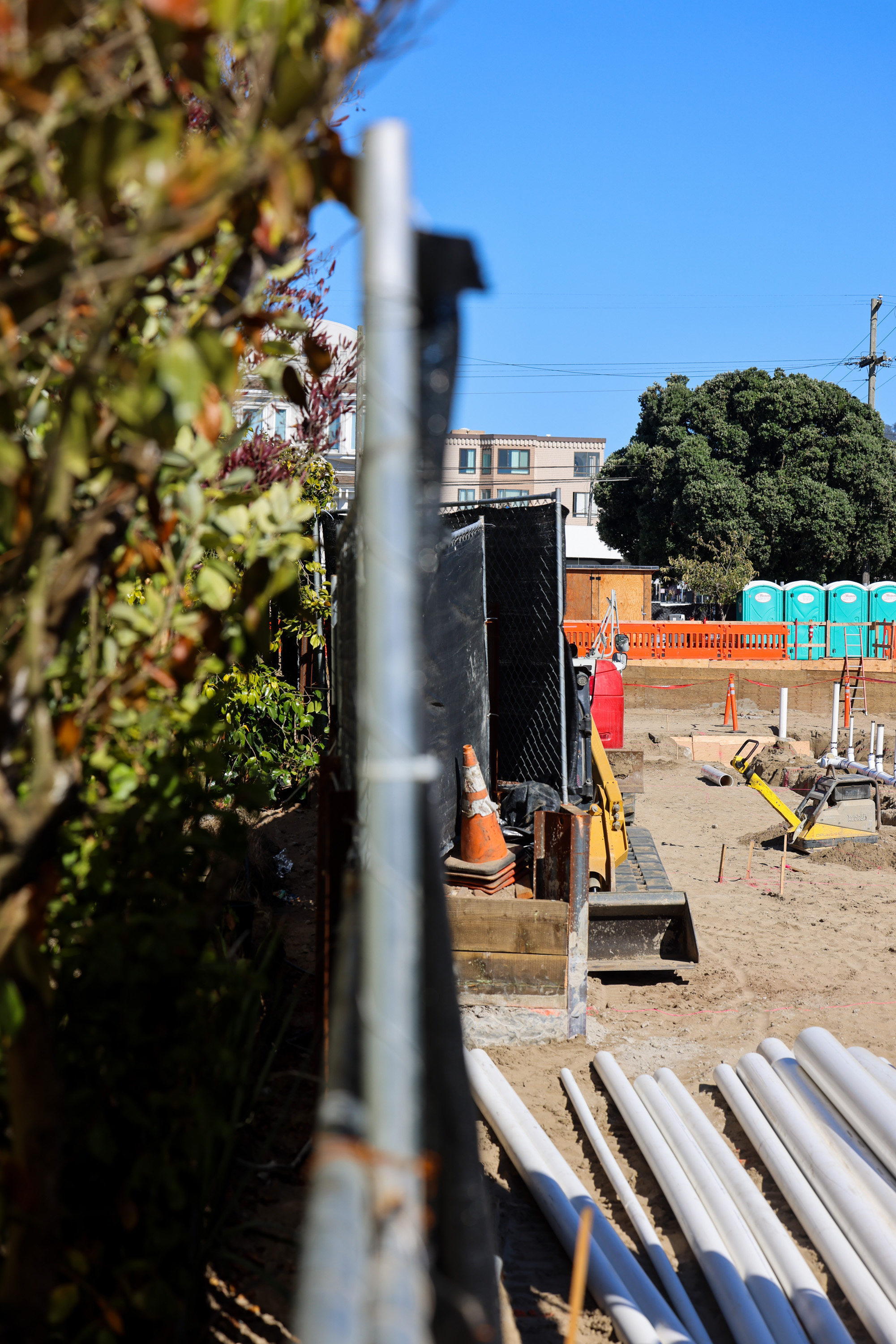 The image shows a construction site with machinery, pipes, and cones. There's a building in the background and portable toilets near a large tree under a clear blue sky.