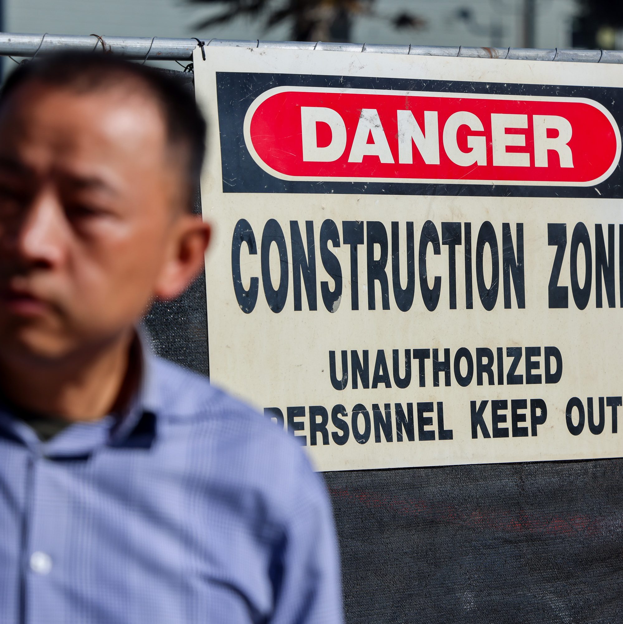 A man in a checkered shirt stands near a &quot;DANGER: CONSTRUCTION ZONE&quot; sign that warns unauthorized personnel to keep out.