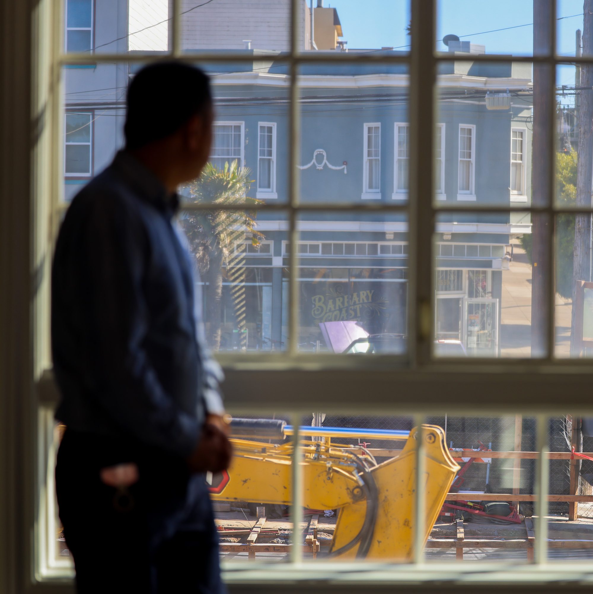 A person is looking out of a window at a construction site with machinery. Across the street, there's a blue building with a sign that reads &quot;Barbary Coast.&quot;