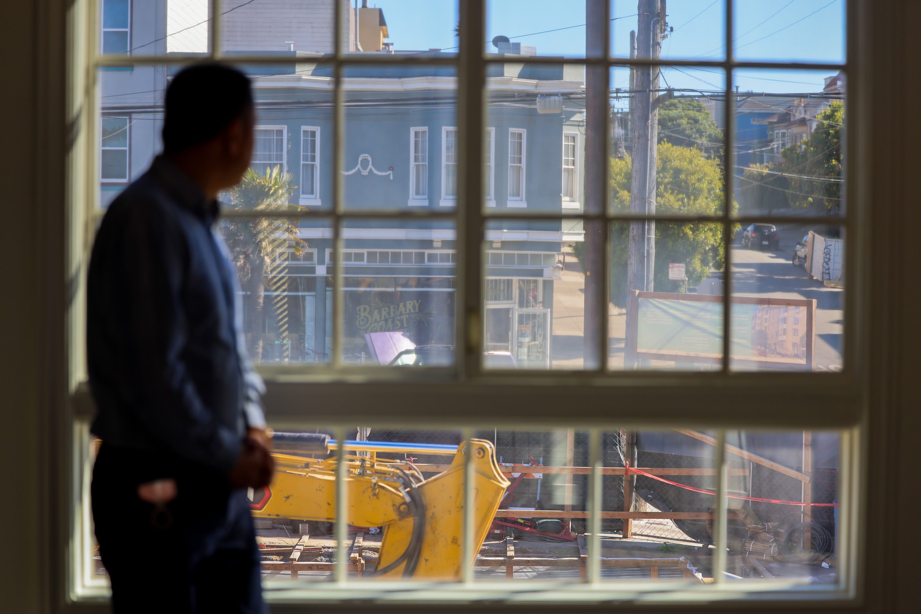 A person stands inside, gazing out a window at a street scene with construction equipment and buildings, one labeled "Barbary Coast," under a clear blue sky.