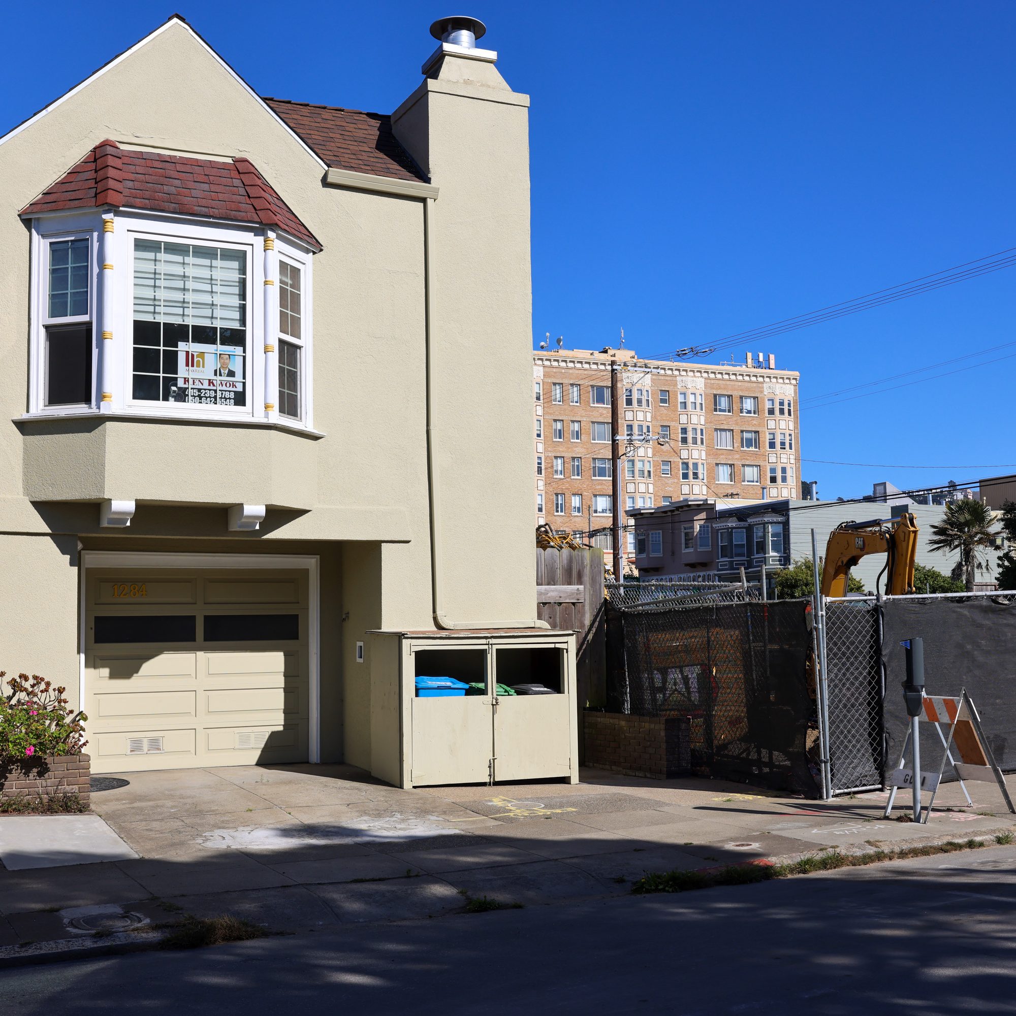 A beige house with a bay window and a garage sits next to a construction site with an excavator, against a backdrop of an old apartment building and clear blue sky.