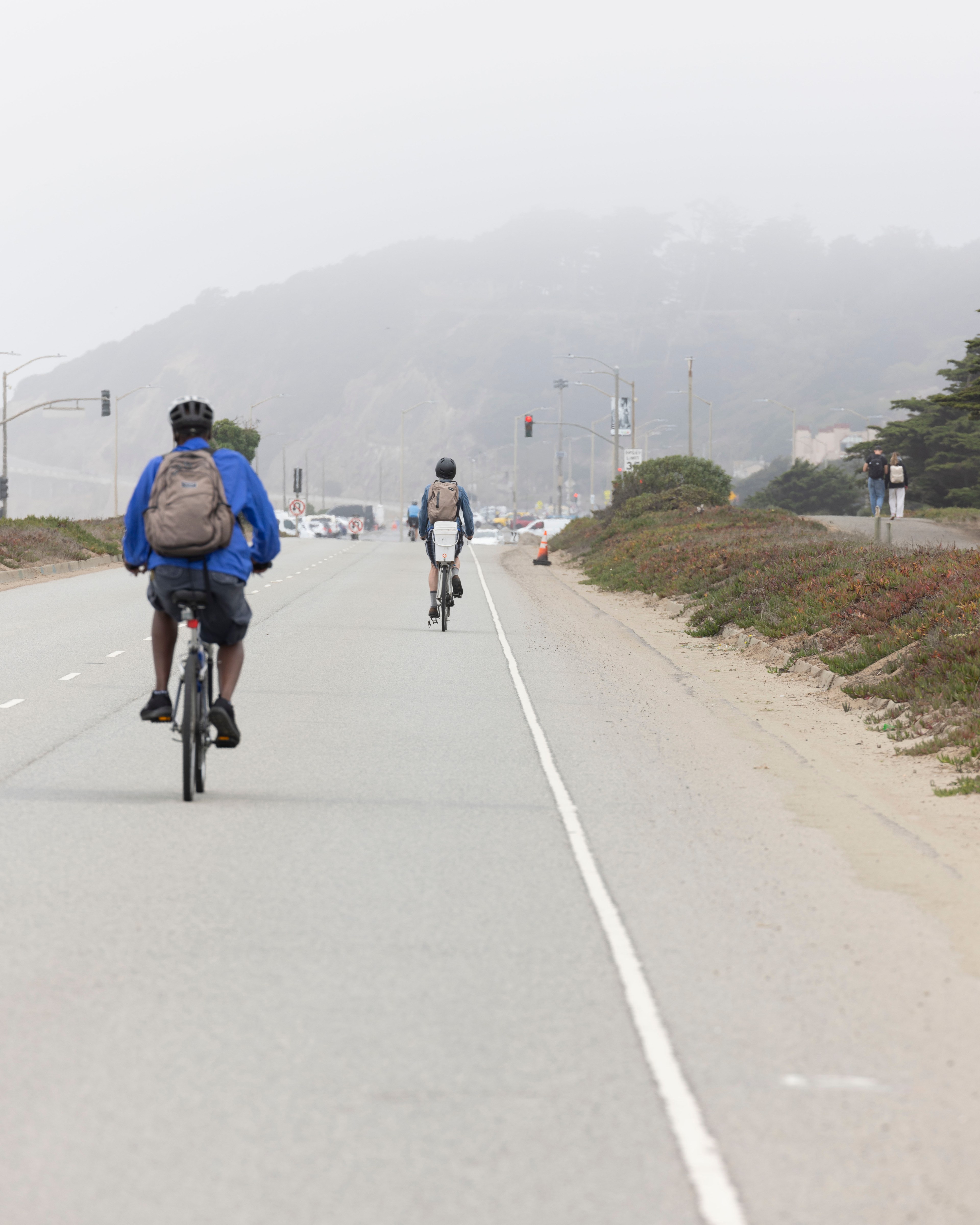 The image shows two cyclists riding on an empty road on a foggy day, with minimal traffic, wearing backpacks and helmets. A landscape with hills and traffic lights is visible ahead.